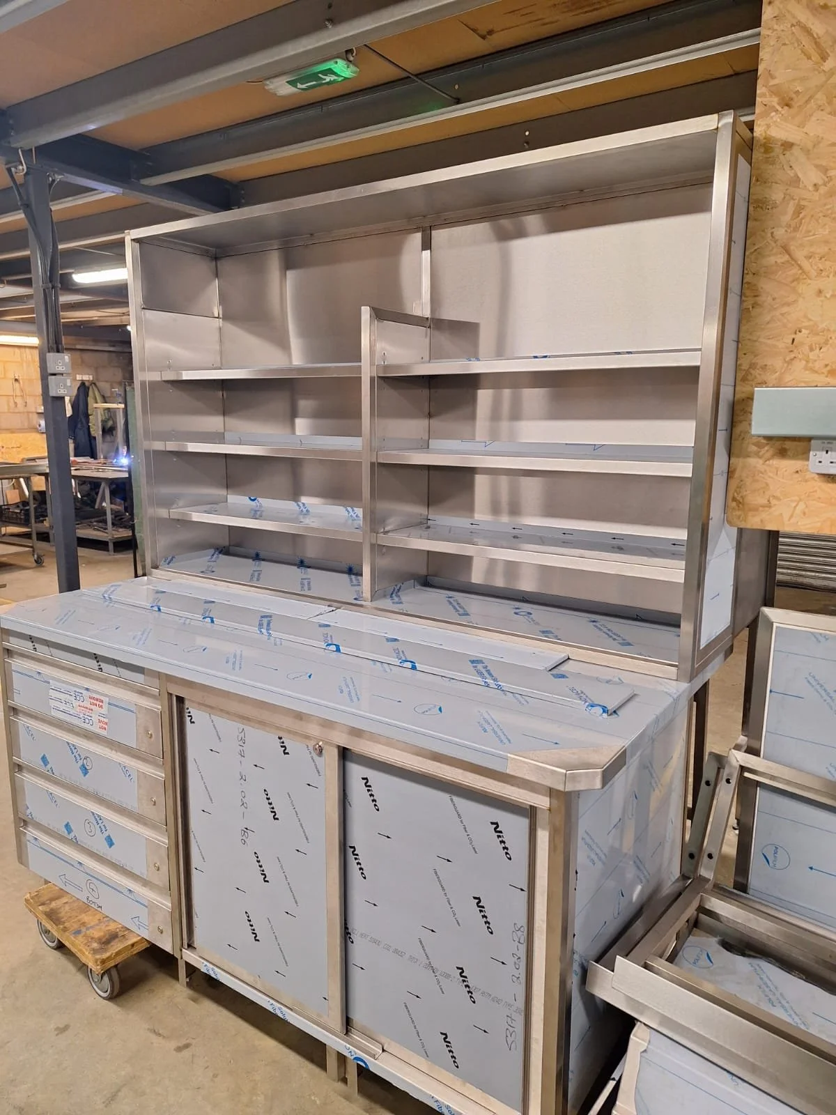 Stainless steel commercial kitchen shelving unit with multiple shelves and drawers, partially covered with protective film, in a warehouse or industrial setting.