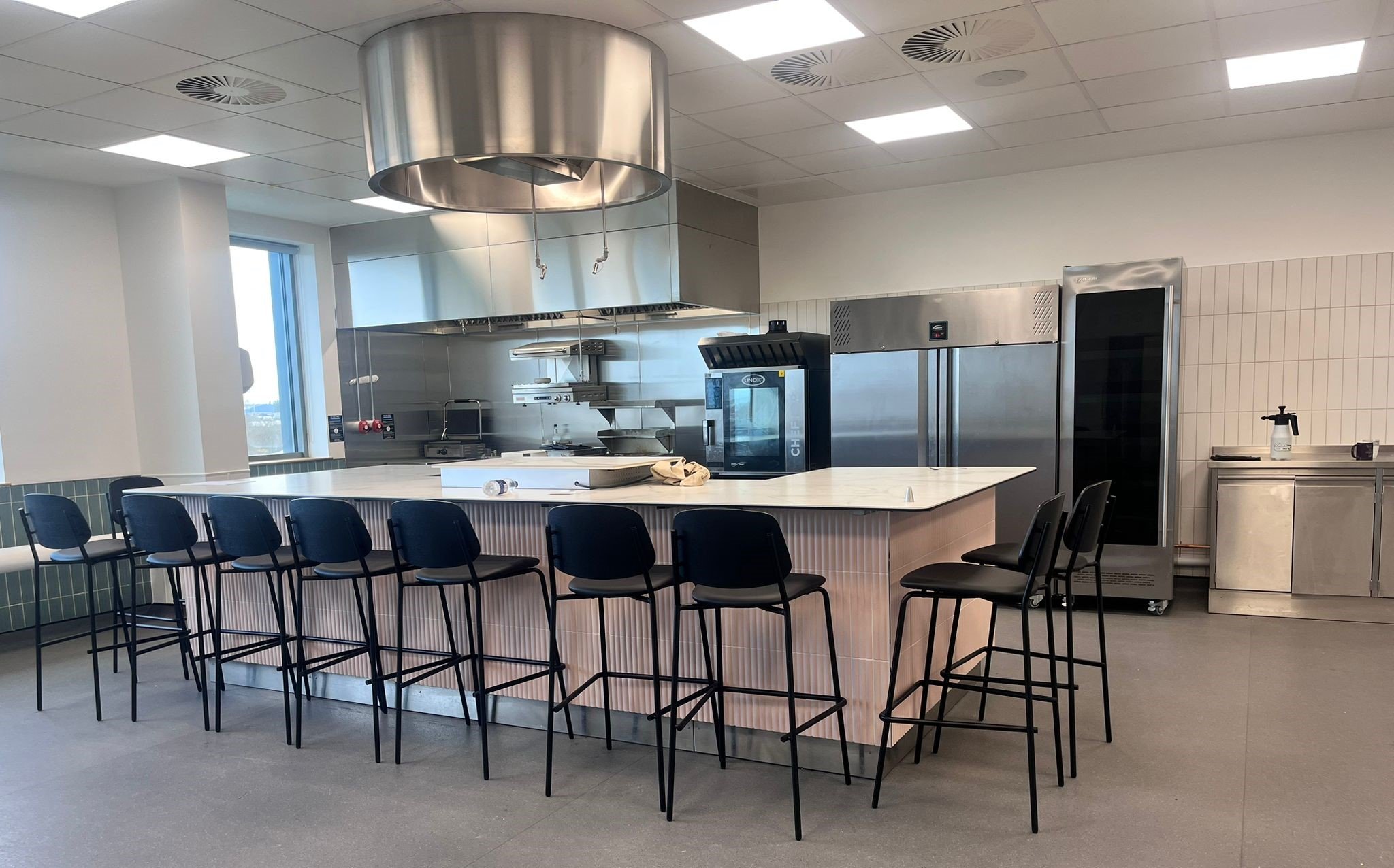 Modern kitchen with a long white counter, black chairs, stainless steel appliances, and a large metallic exhaust hood overhead.