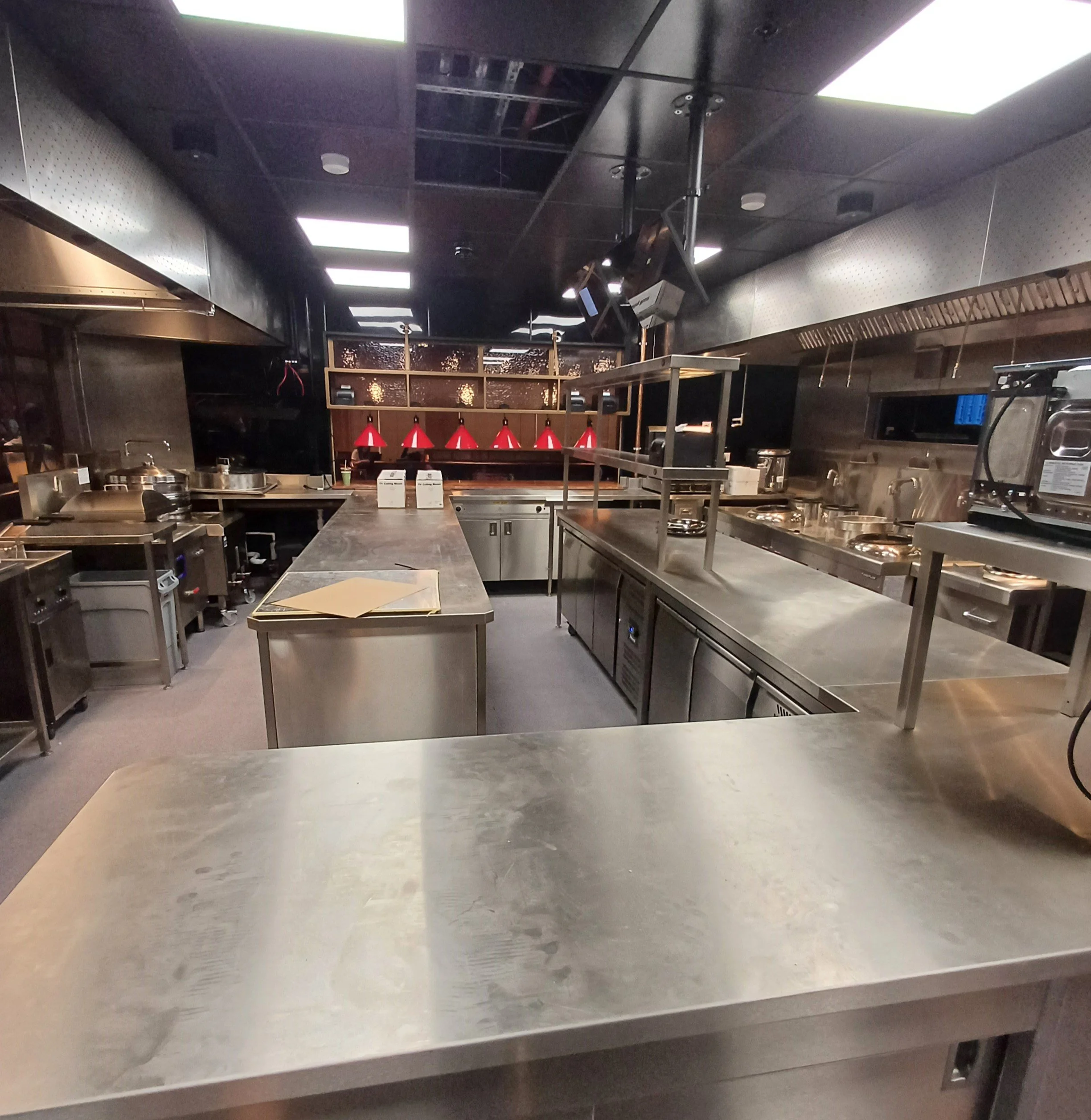 Empty commercial kitchen with stainless steel counters, ovens, and red pendant lights in the background.