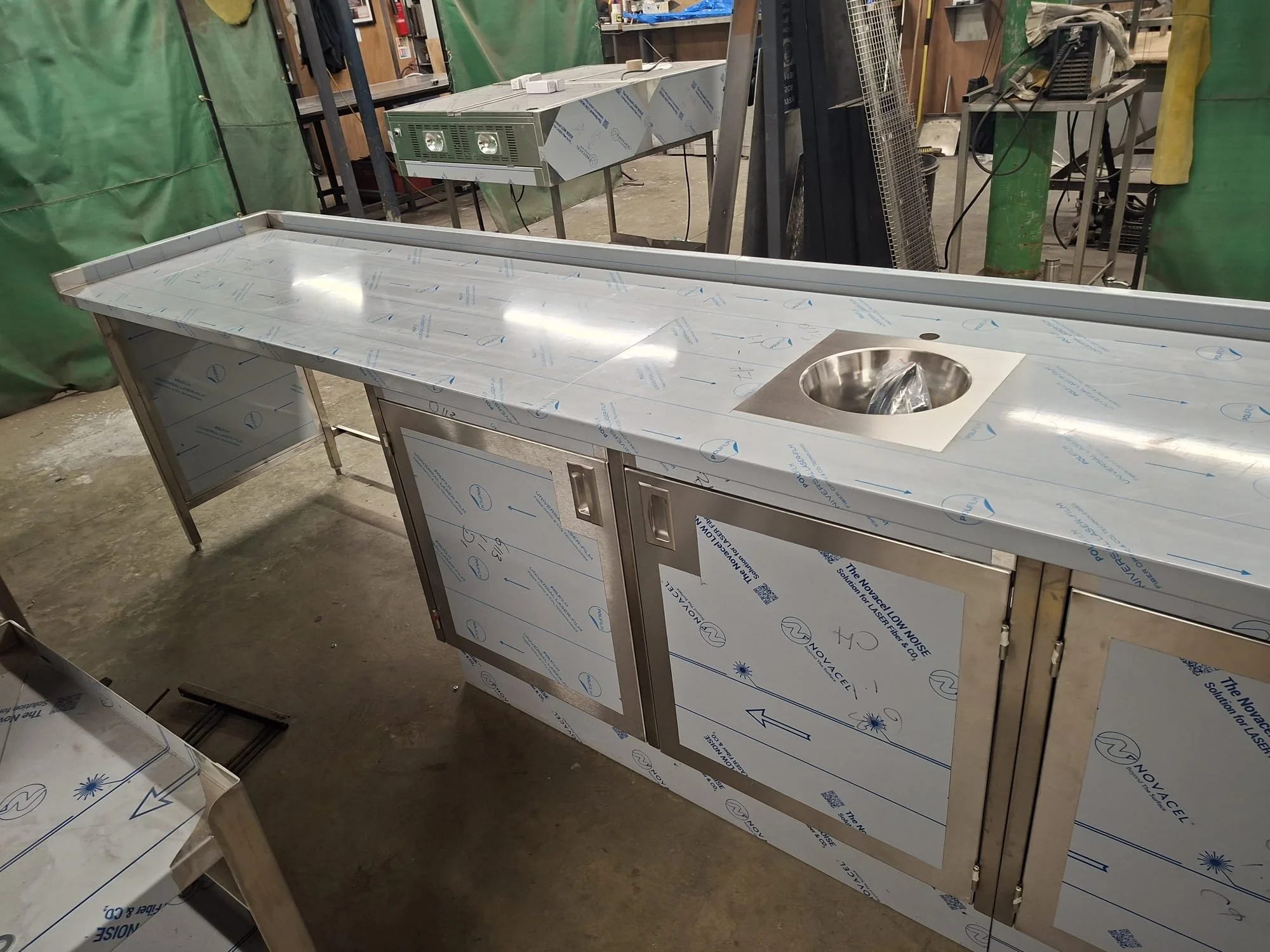 Stainless steel commercial kitchen countertop with integrated sink in a workshop or factory setting, surrounded by green safety barriers, equipment, and tools, with protective film still attached.