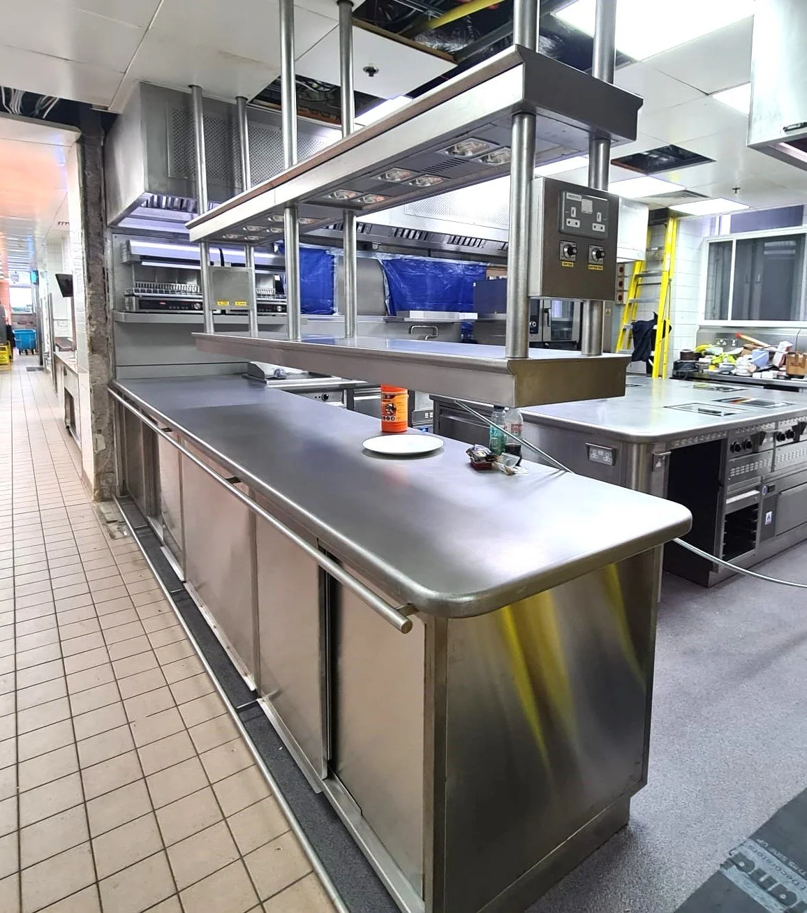Empty stainless steel commercial kitchen countertop with a plate, soda bottle, and some small items on it, surrounded by industrial cooking appliances and shelves.