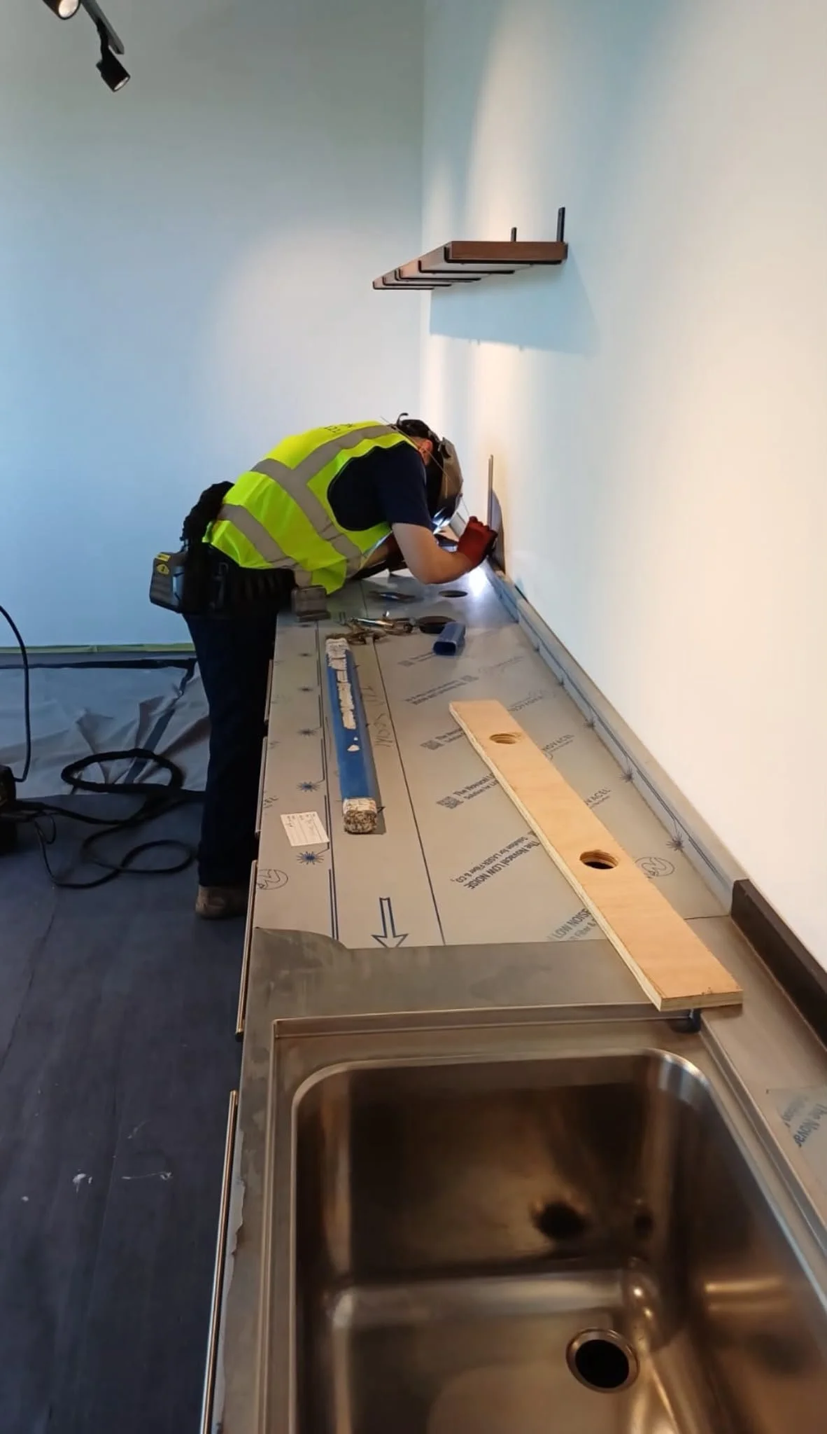 A worker in a yellow safety vest and red gloves is installing or working on a stainless steel sink in a room with white walls. The worker is bent over a counter with tools and materials around. The photo is taken from above, showing the sink and part