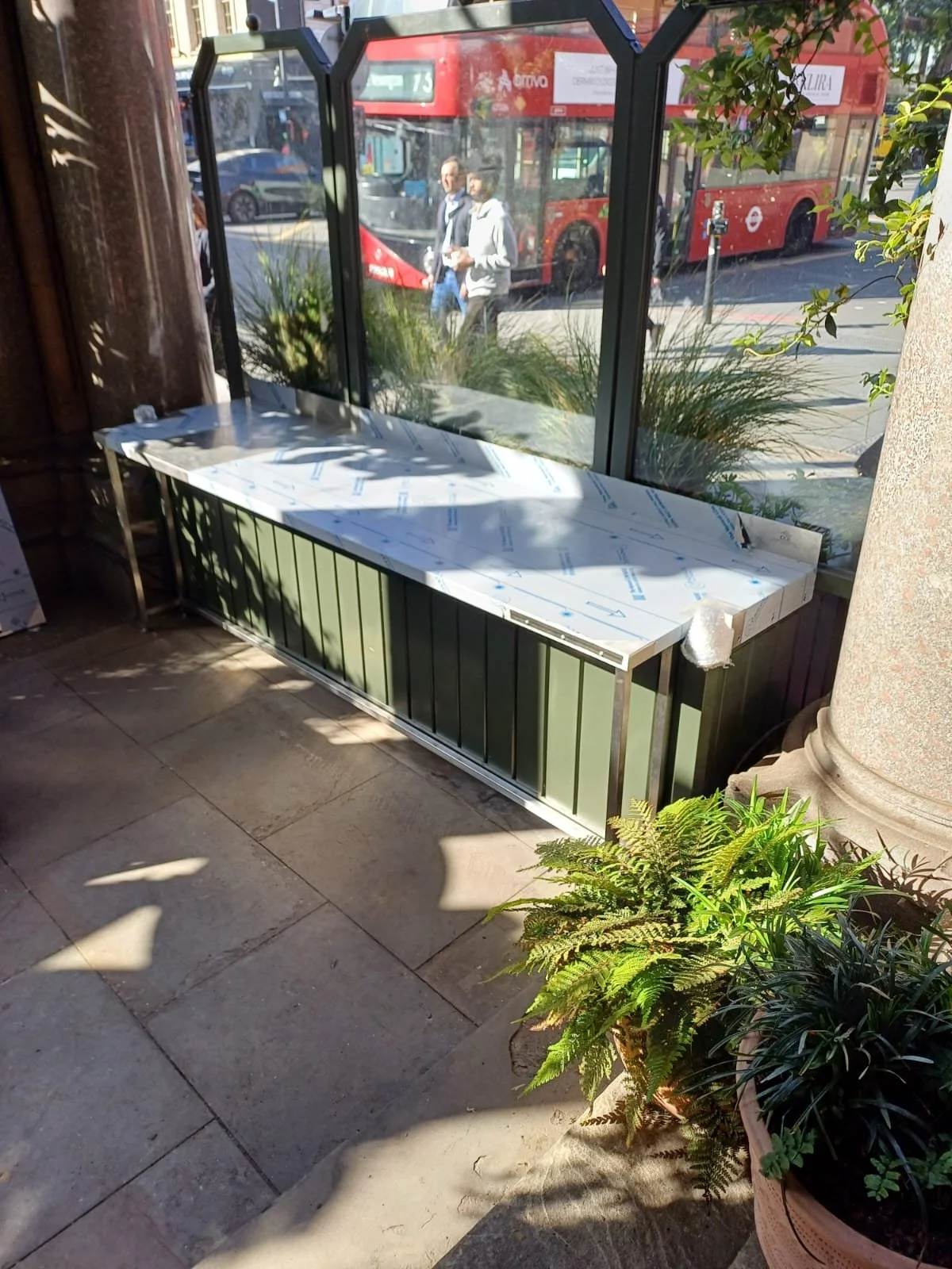 Empty green metal bench next to a large window with plants outside and a busy city street with a red double-decker bus passing by.
