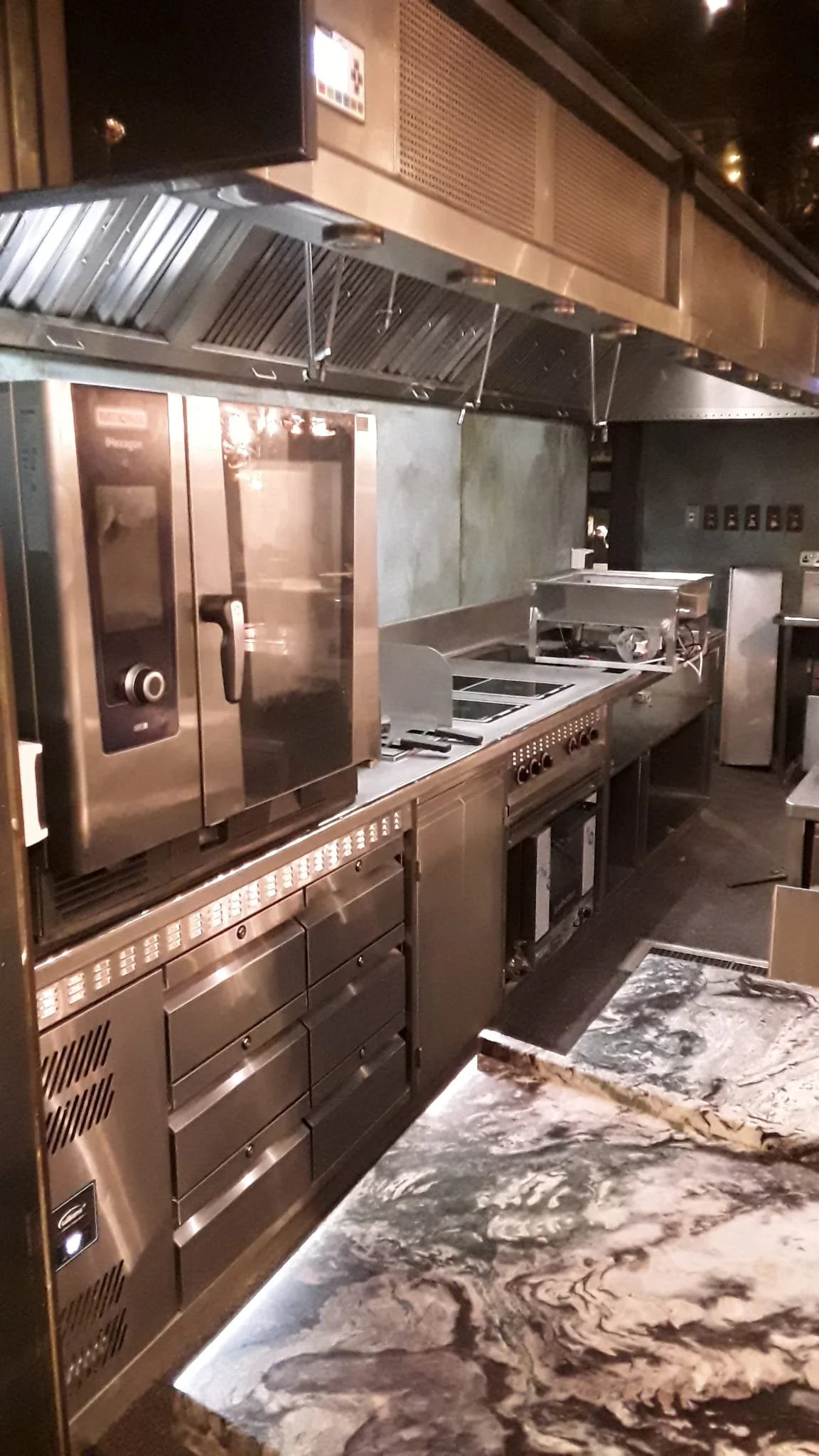 Commercial kitchen with stainless steel appliances, including an oven, stove, and storage drawers, and a marble countertop with a black and white swirling pattern.