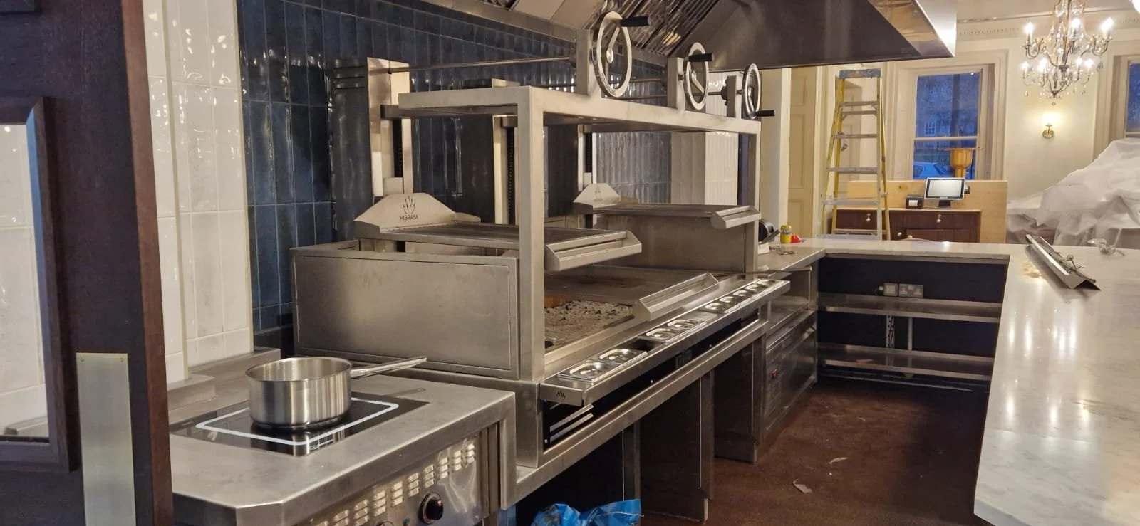 Commercial kitchen with stainless steel appliances, a stovetop with a pot, and various cooking equipment. In the background, there is a ladder, a wooden cabinet, and a chandelier.
