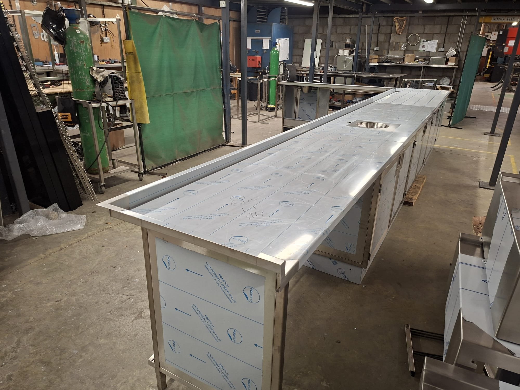 A stainless steel kitchen counter in a workshop, with a hole for a sink, surrounded by tools, tanks, and industrial equipment in the background.