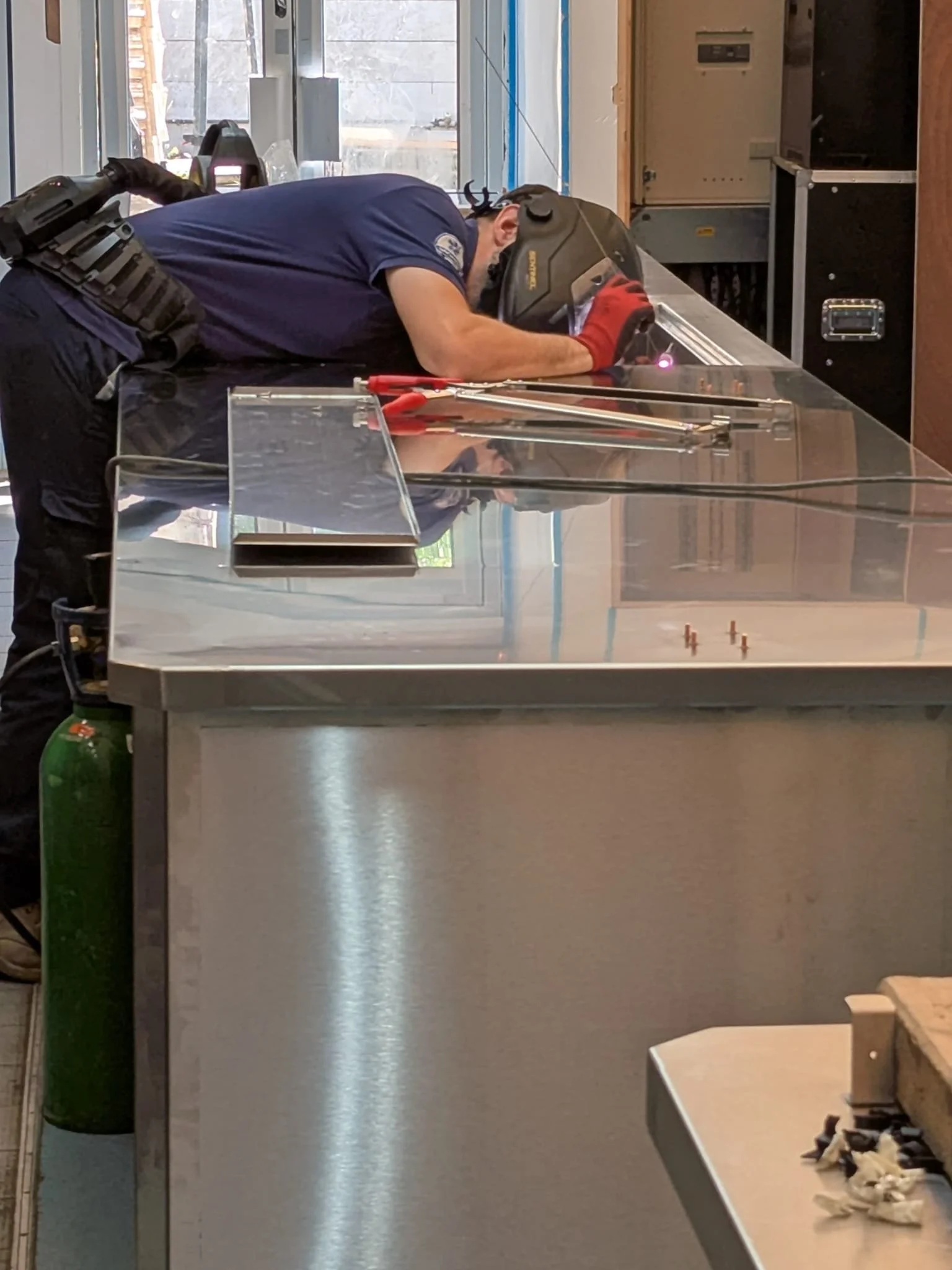 A person welding on a metal surface indoors, wearing a helmet and red gloves, with tools and equipment nearby.
