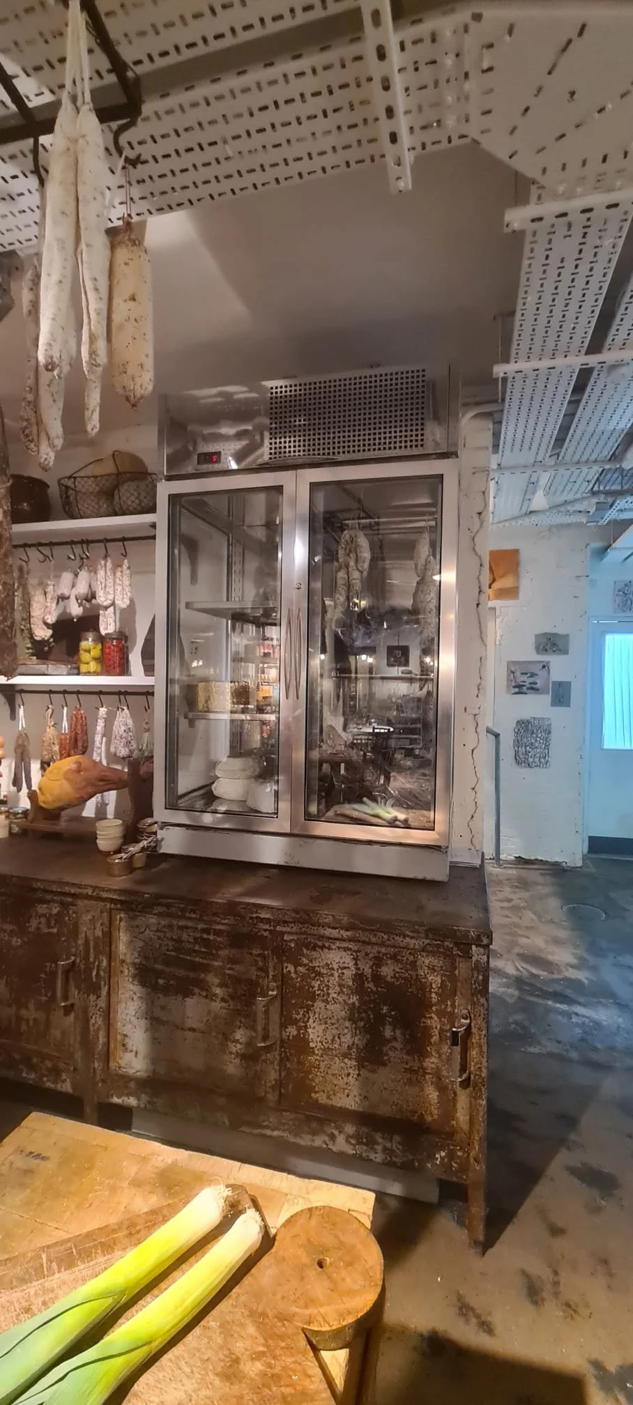 A rustic kitchen with a vintage wooden cabinet, glass refrigerator with hanging cured sausages, shelves with spices and hanging meat, and a cutting board with leeks on a table.