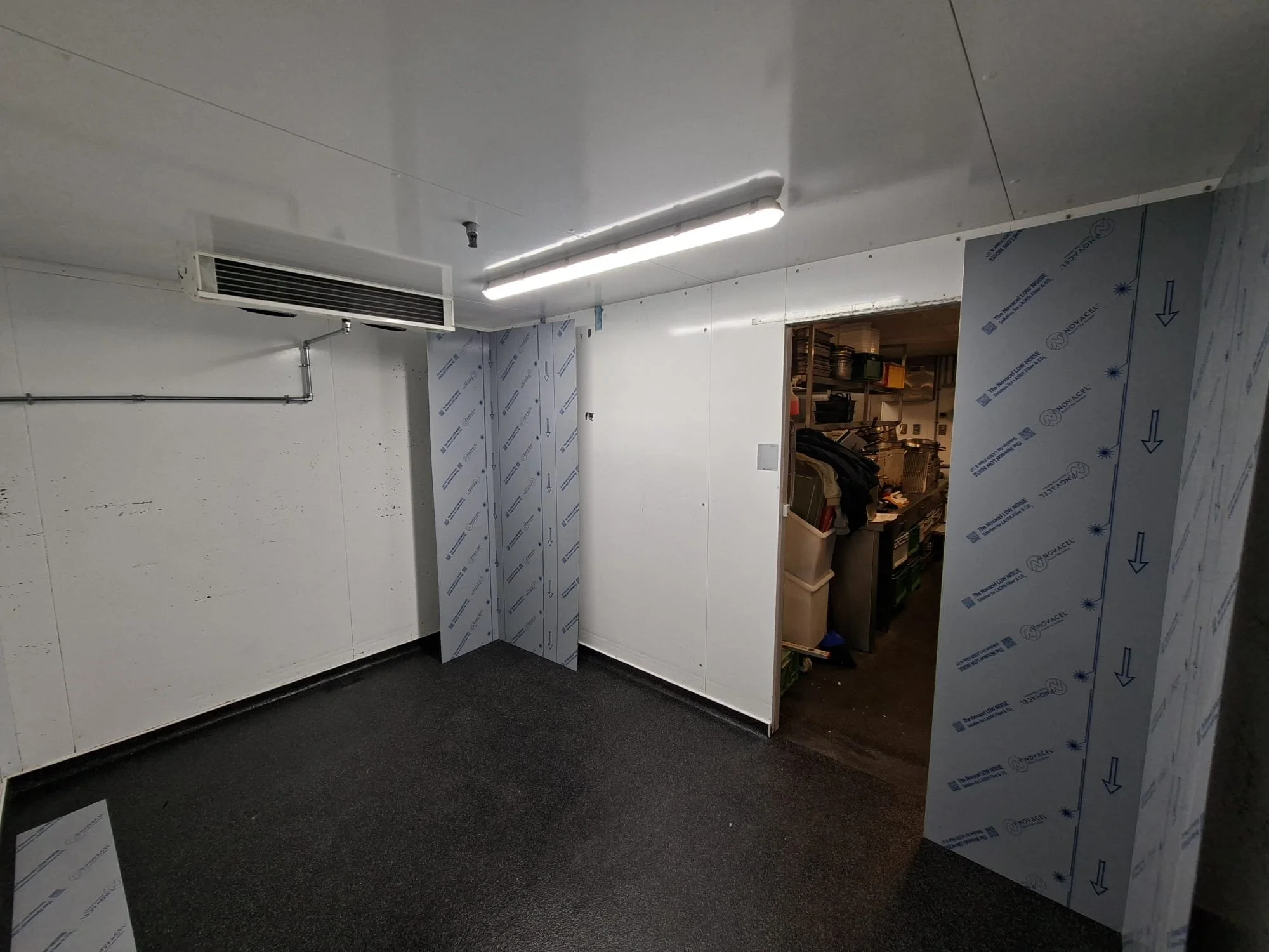 An unfinished room with white walls, a black carpeted floor, rectangular fluorescent lighting, an air duct, and an opening leading to a storage room filled with shelves and containers.