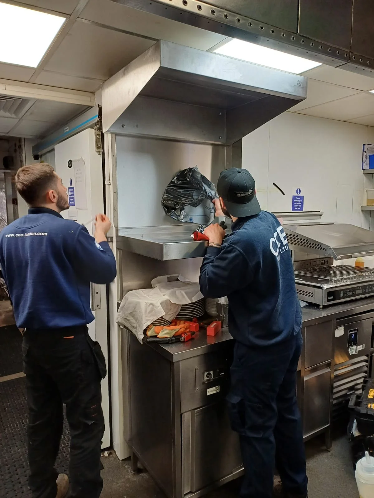 Two workers in a kitchen, one opening a large industrial oven, the other standing nearby, with tools and equipment on the counter.