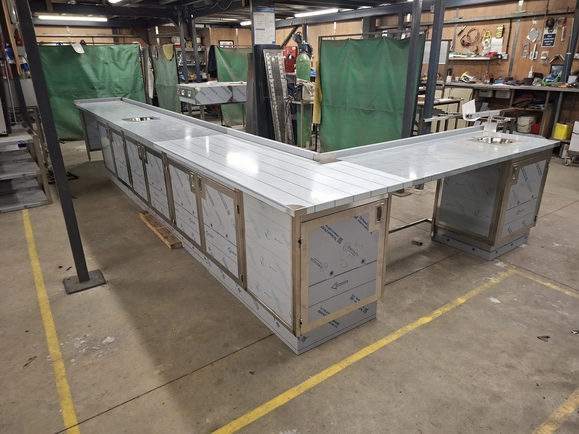 A stainless steel kitchen island with storage cabinets underneath and a cutout in the countertop, situated in an industrial workshop. The workshop background includes green curtains, tools, gas tank, and workbenches.