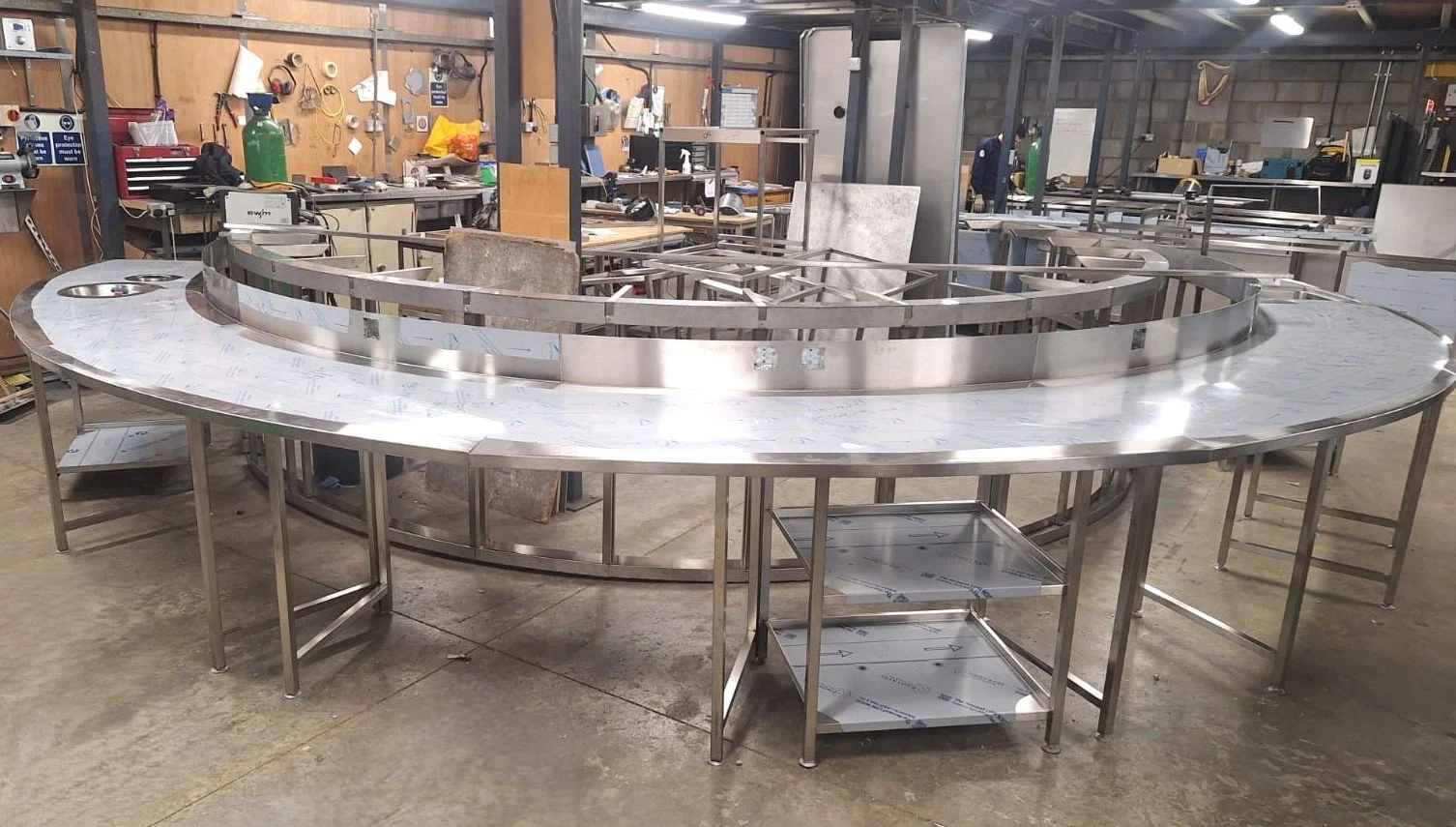 Stainless steel curved kitchen or bar counter in a workshop or industrial setting with tools and equipment in the background.