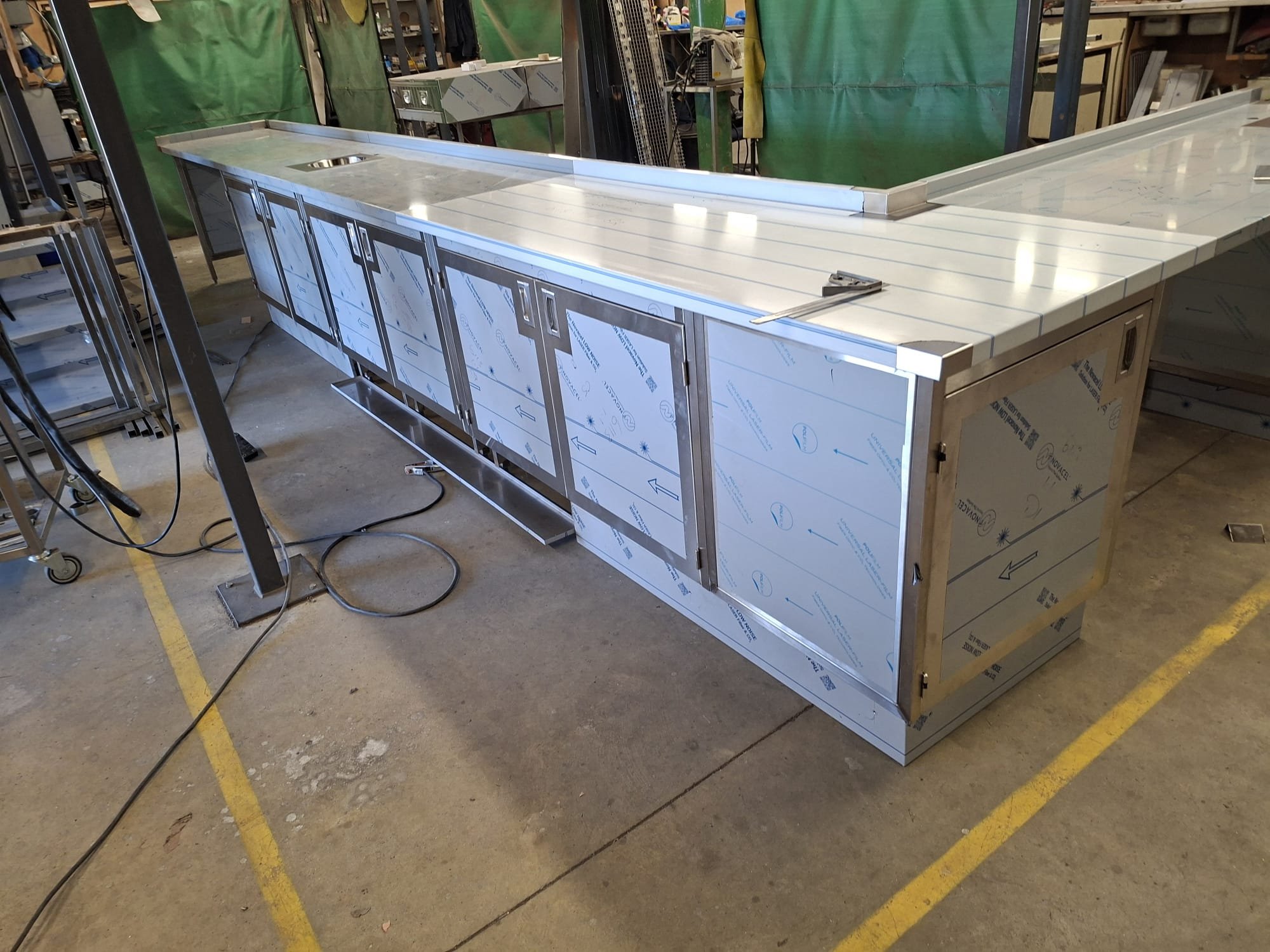 A large industrial stainless steel work station in a workshop or fabrication area, with tools and other equipment nearby.