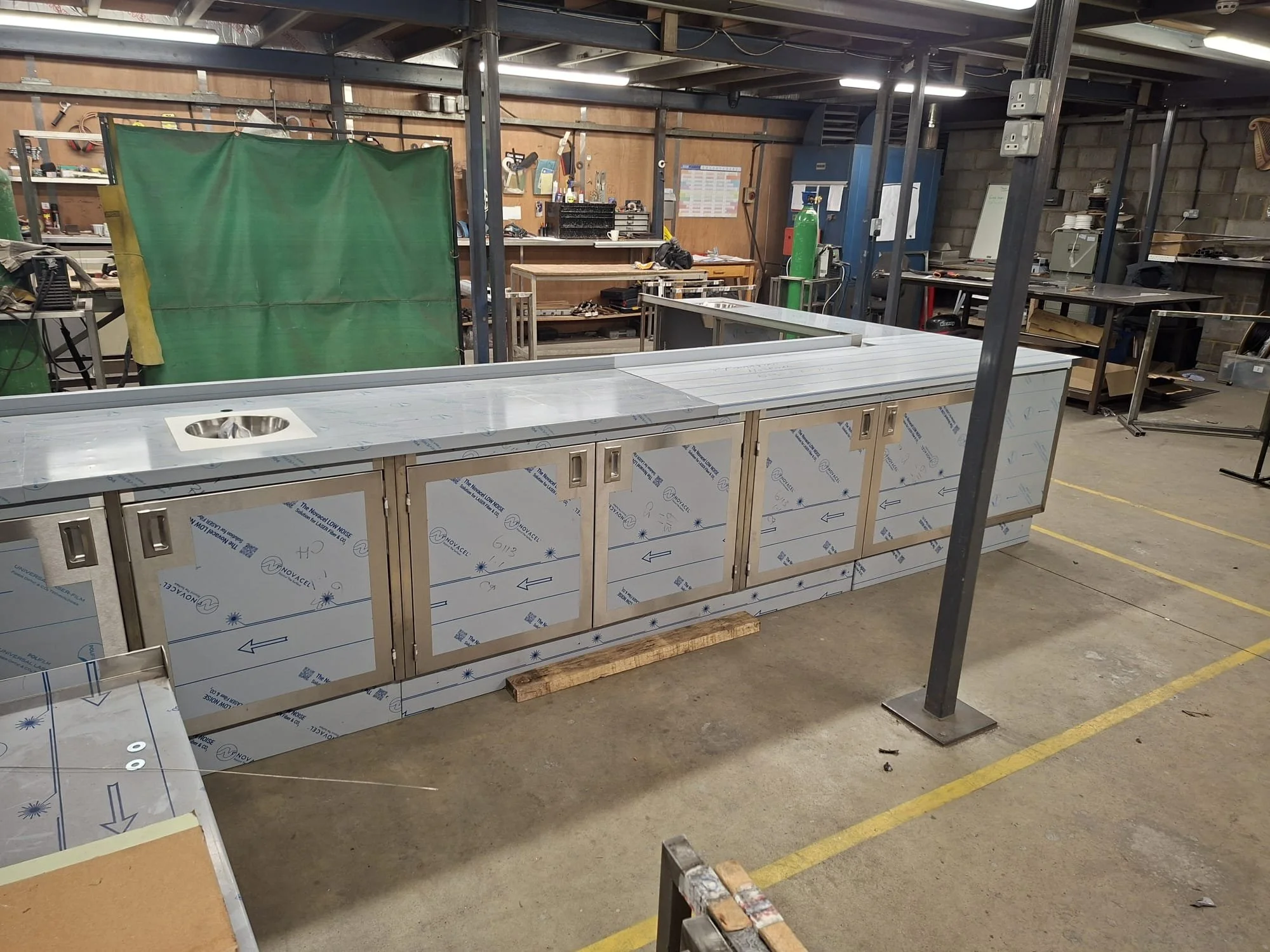 Interior of a workshop with metal cabinets, tools, and equipment. A partially assembled metal structure is in the foreground. The space has a concrete floor with yellow lines and a brick wall at the back.