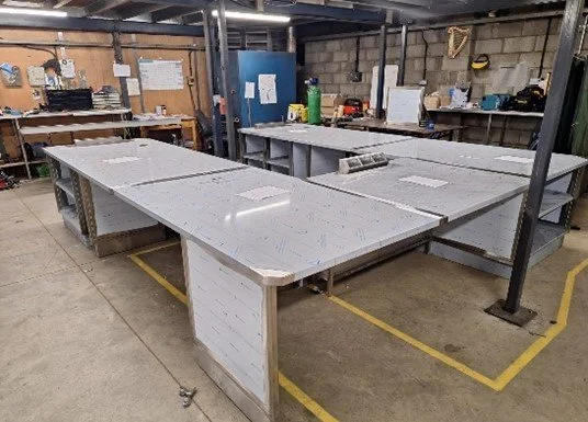 White workbenches in a workshop or industrial space with tools and supplies on shelves in the background.