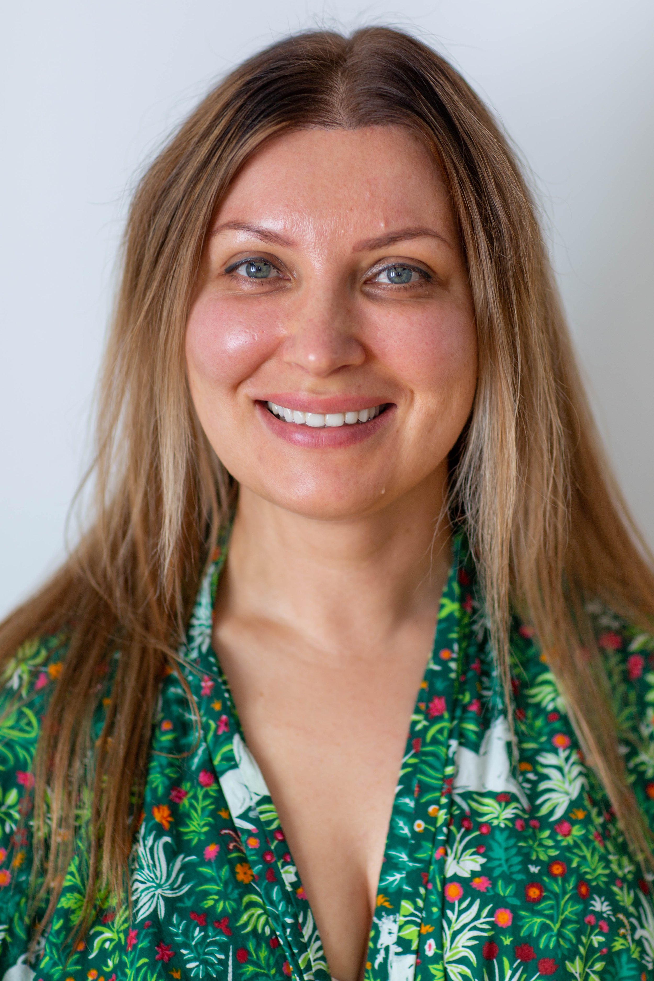Close-up of a woman with long light brown hair, blue eyes, and a friendly smile, wearing a green floral shirt against a plain white background.