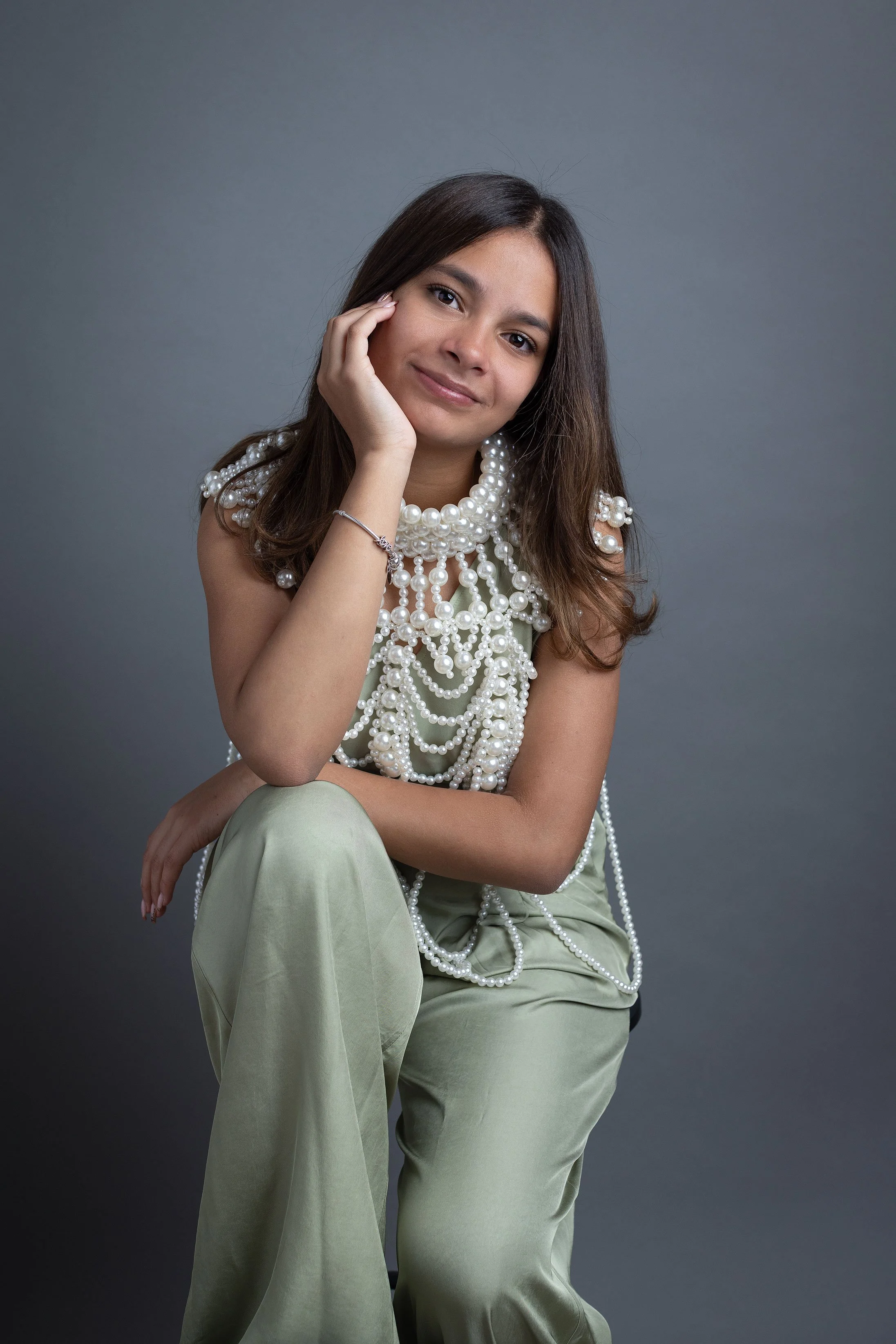 A young woman with long brown hair wearing a pearl necklace and bracelet, sitting against a gray background, smiling softly, with her hand resting on her face posing for Monika M. Kubiak, a  professional photographer for women in Leonberg and the Stu
