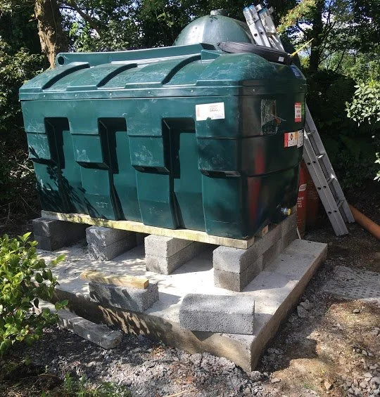 Large green obunded oil tank on concrete blocks, with ladder and trees in the background.