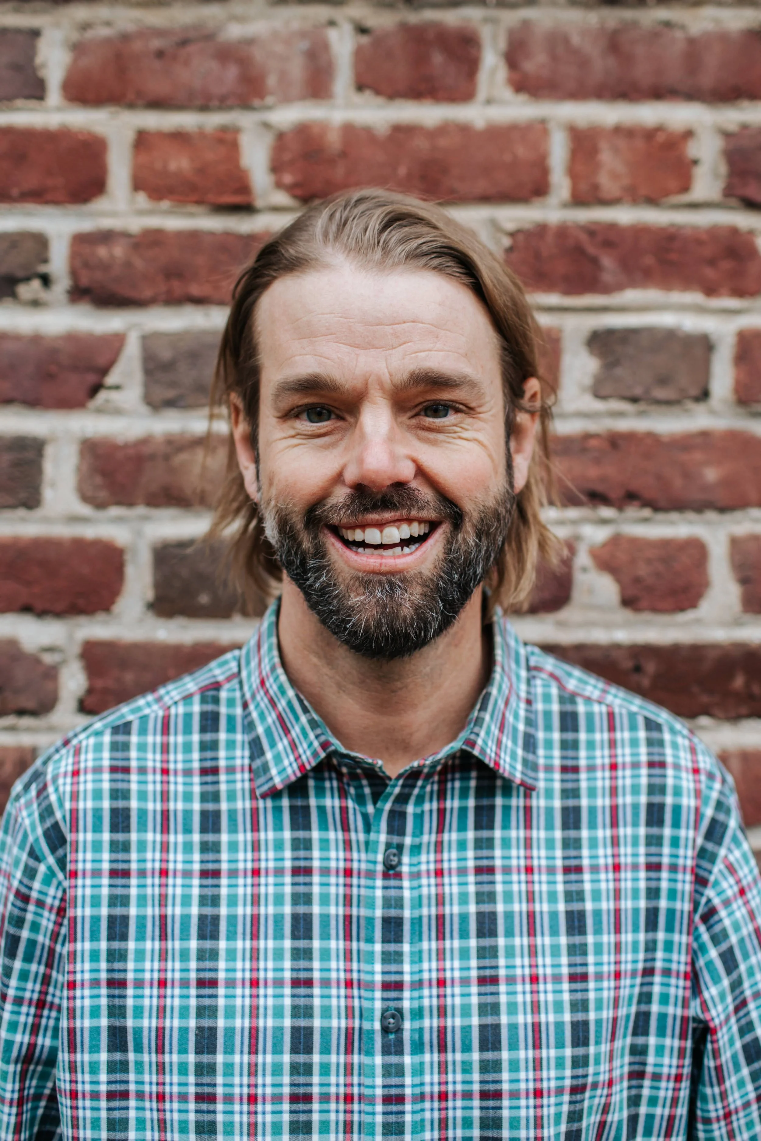 A man with long hair and a beard smiling, standing in front of a brick wall, wearing a plaid shirt.