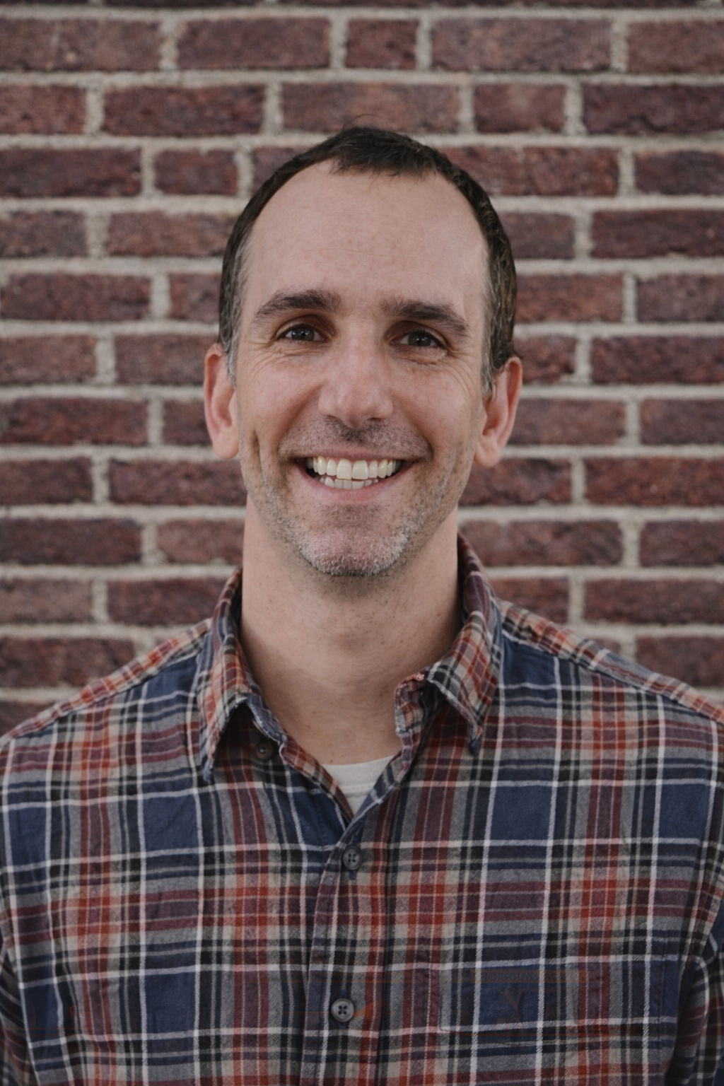 Portrait of a smiling man in front of a brick wall, wearing a plaid shirt.