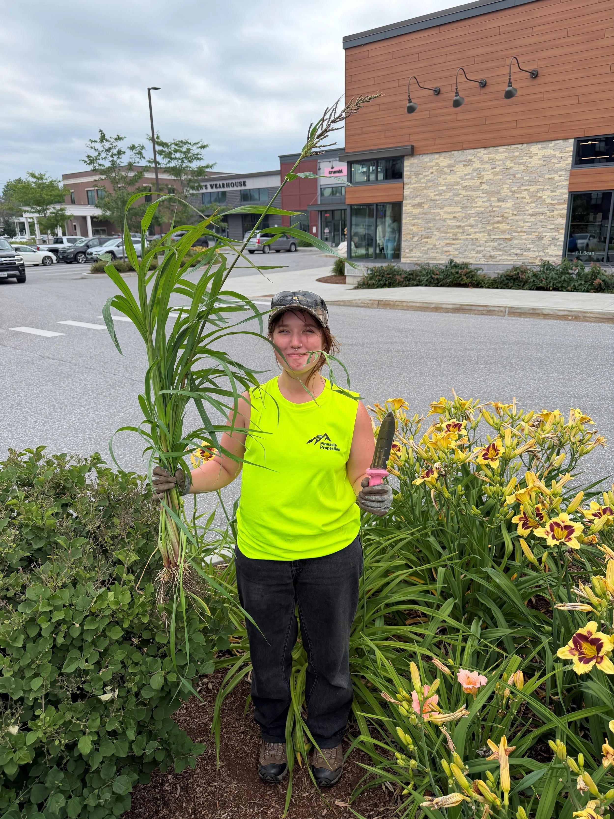 A smiling girl in a bright yellow tank top holding up a weed and a soil knife.