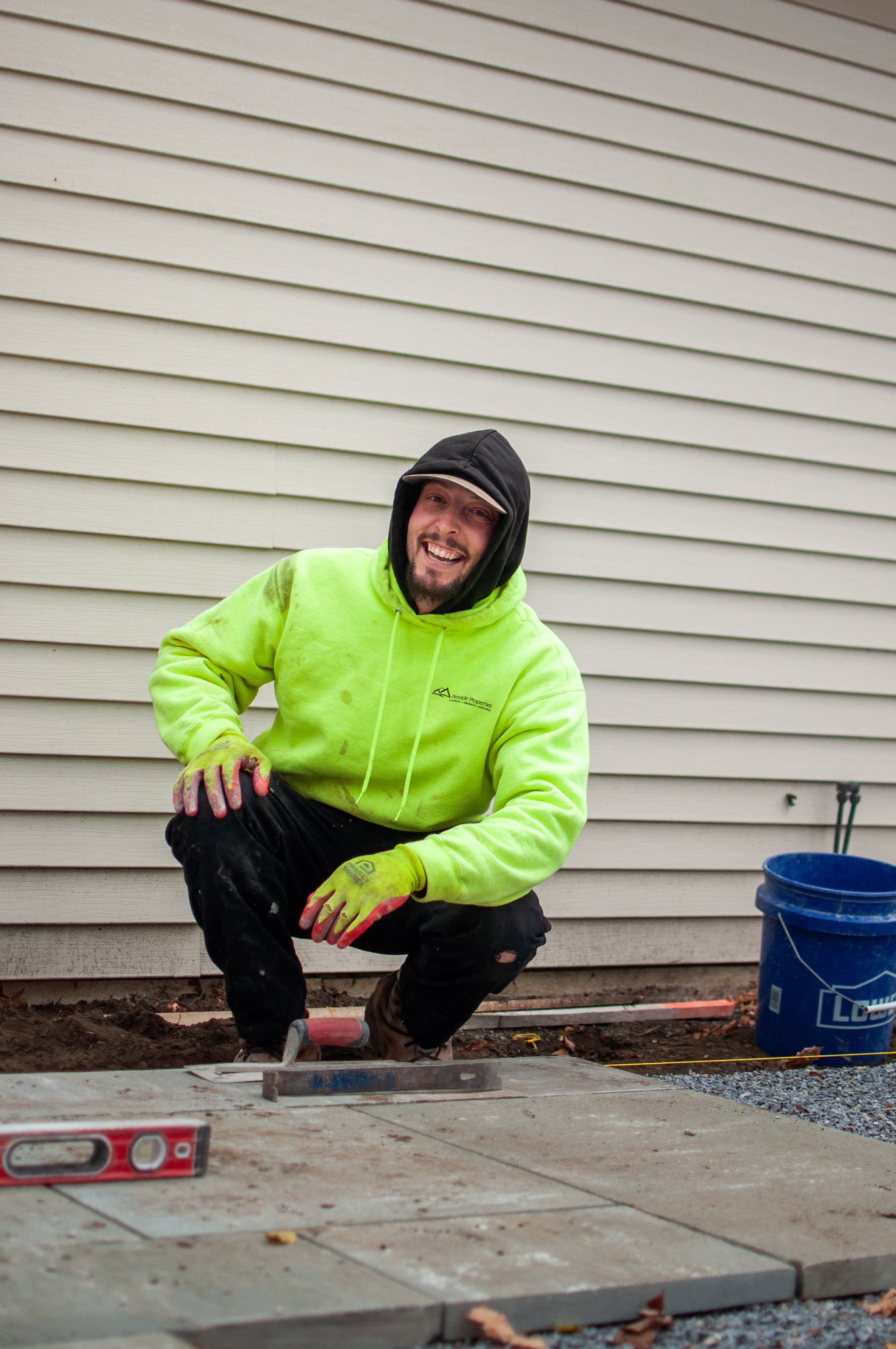 A person in a neon yellow shirt and cap holding a tool stands in front of a brick building.