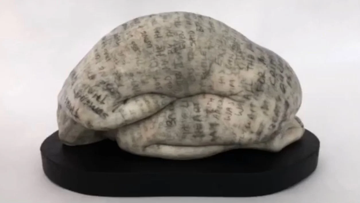 Sculpture of a person completely curled up with their head between their legs - The skin is tattooed with black text - The person is seated on a black pedestal - Robin Amora - Verduyn