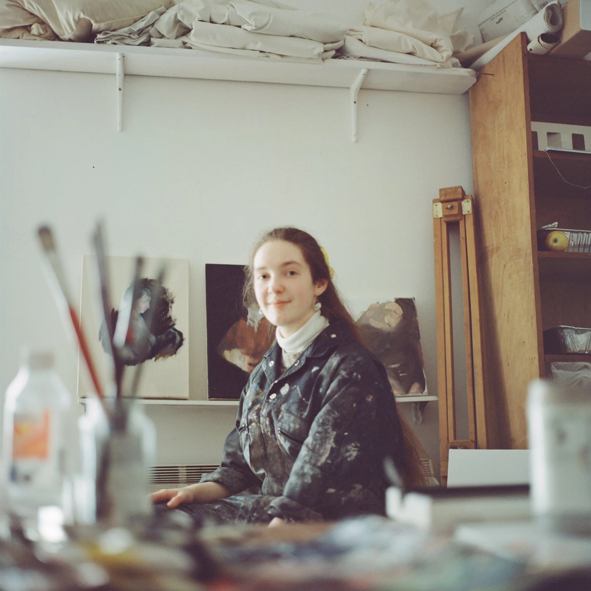Portrait of the artist Sian Costello sitting in her own studio. She is looking directly at the camera, with the foreground showcasing her brushes.