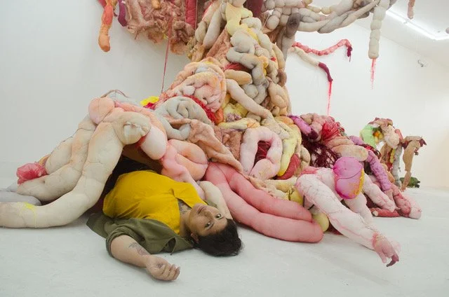 Maria Abaddon lying on the ground beneath an installation of primarily white and pink sausage-shaped stuffed objects. Within the installation, a stuffed figure appears to be emerging.