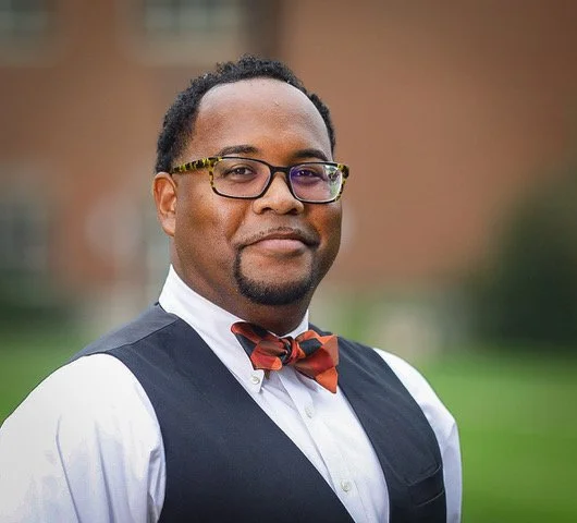 Man wearing glasses, white shirt, black vest, and red bow tie standing outdoors in front of a blurred background.