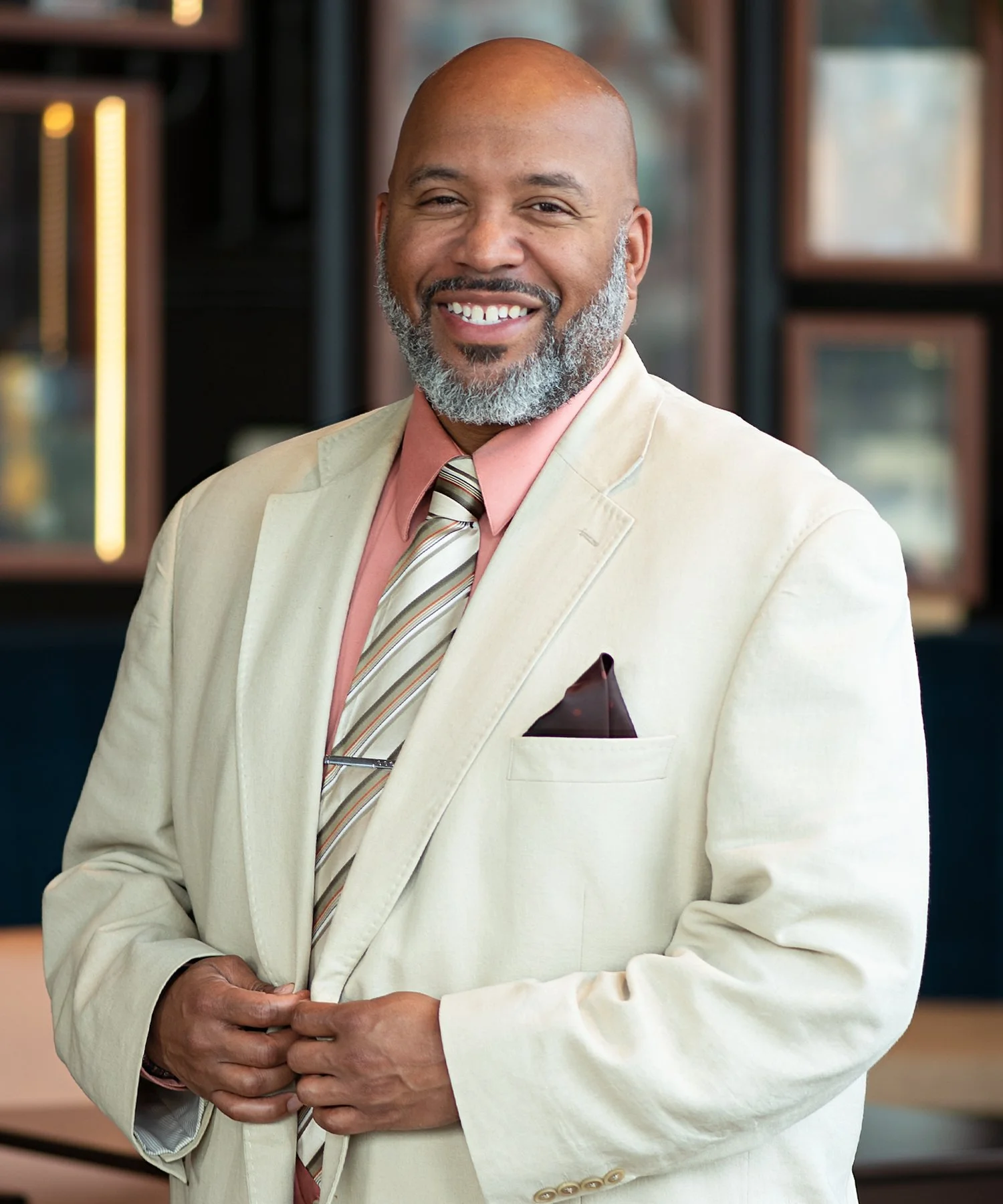 A professional man smiling, with a bald head and full beard wearing a beige suit, pink shirt, and patterned tie, looking at the camera.