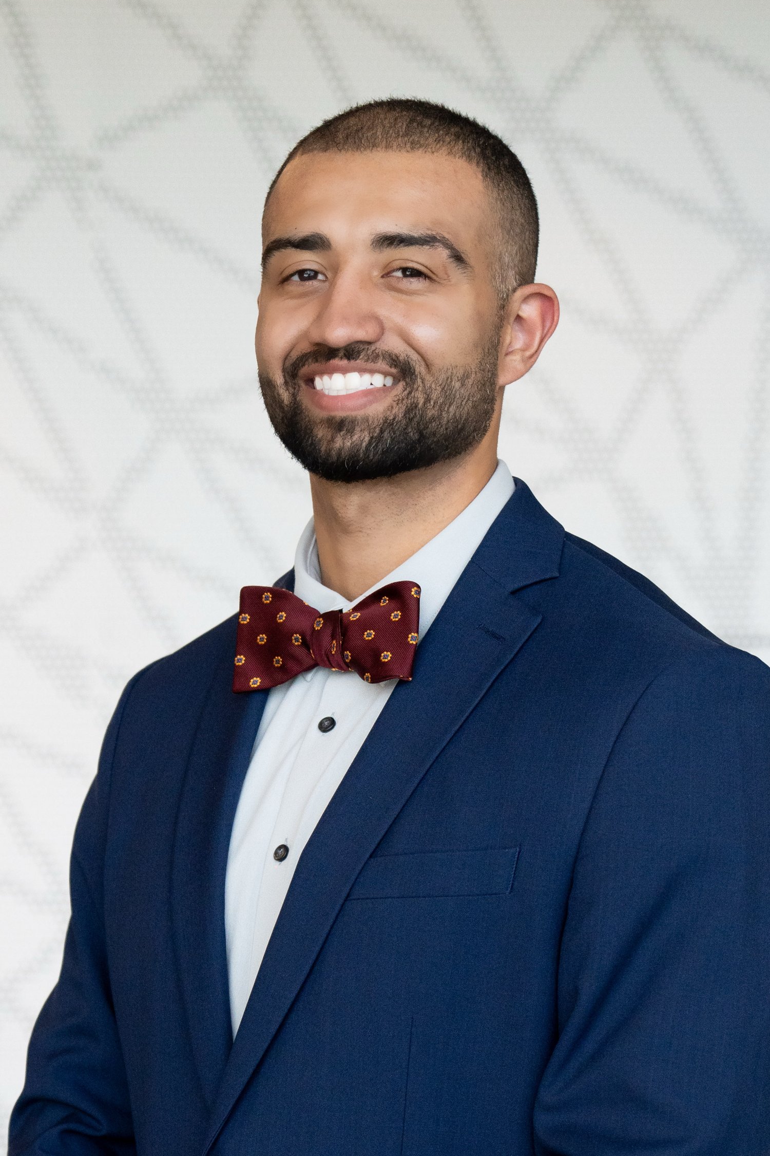Man wearing white shirt, blue blazer, and red bow tie smiling in front of a patterned background.