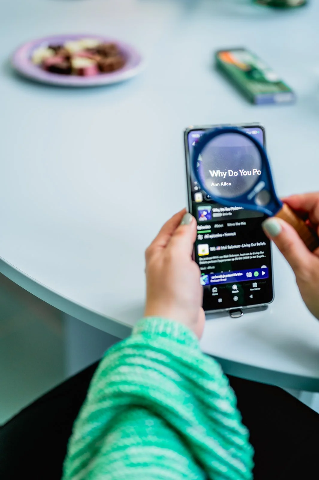 Close-up foto van de telefoon van Ann Alice, op het scherm zie je haar podcast Why Do You Podcast op Spotify, ze kijkt ernaar met een vergrootglas