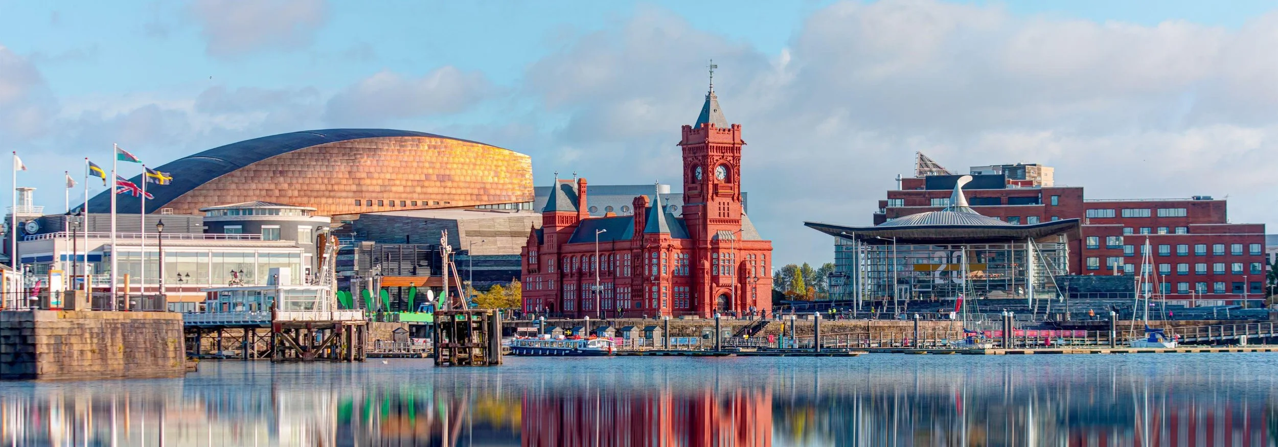 City skyline featuring a red historic building with a clock tower, modern architecture, and flags along the waterfront, with reflections in the water.