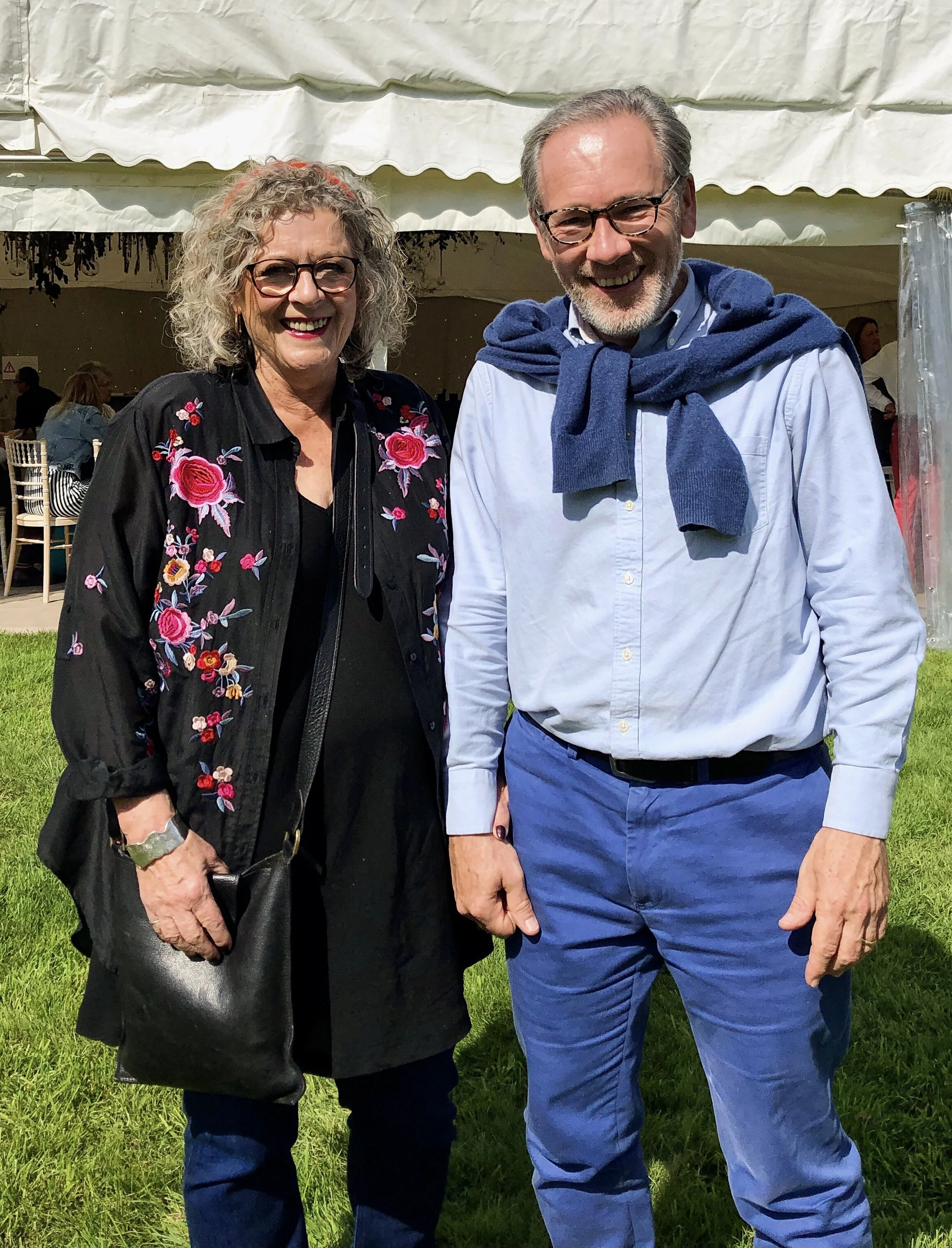 A woman and a man standing outdoors in front of a tent, smiling at the camera. The woman has curly gray hair, glasses, and is wearing a black outfit with a floral embroidered black jacket. The man has gray hair, glasses, a light blue button-up shirt,