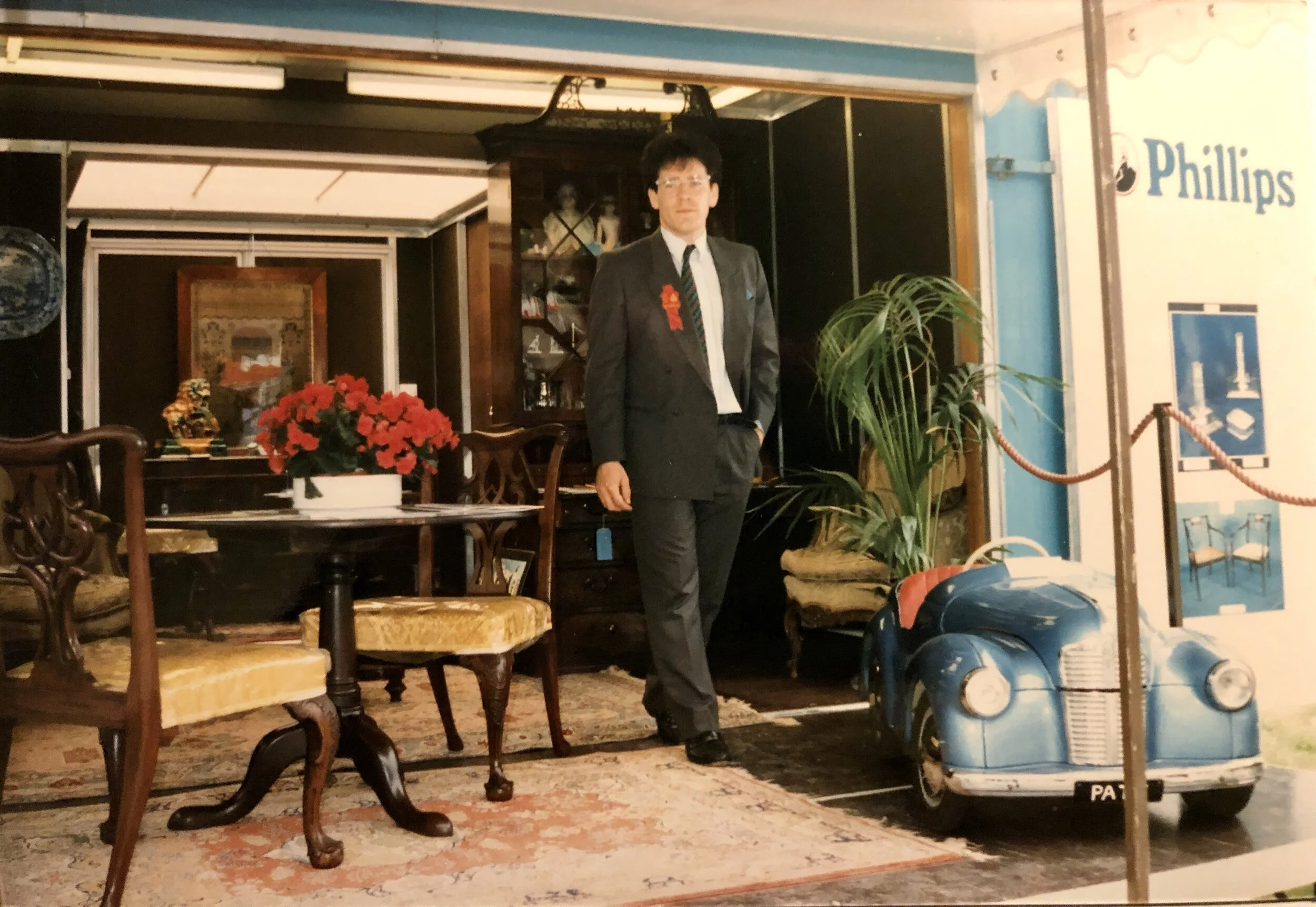 A man in a suit standing inside a vintage furniture shop, with antique chairs, a table with a red flower arrangement, a large potted plant, and a small blue toy car.