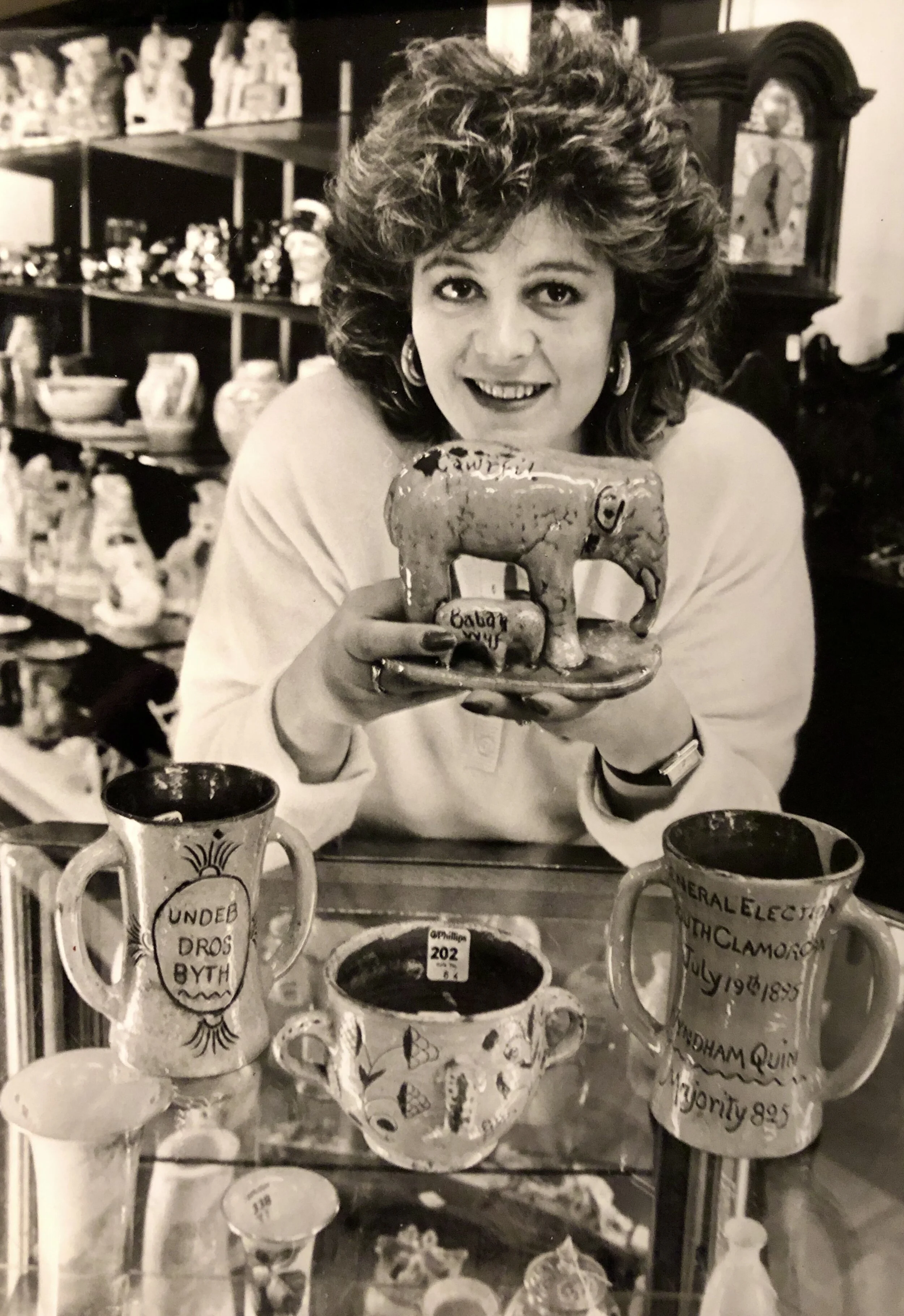 Black and white photo of a woman with curly hair, smiling and holding a small ceramic elephant figurine. She is sitting at a table with decorative pottery and cups in front of her, with shelves full of various ceramic items in the background.