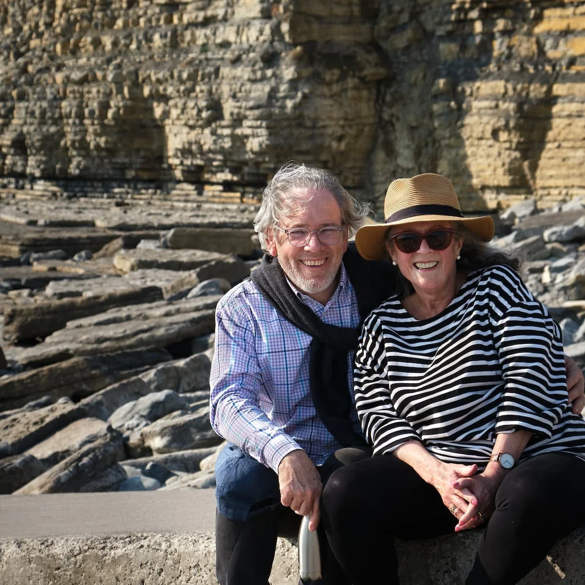 A smiling middle-aged man and woman sitting on a concrete ledge on a rocky beach with layered cliffs in the background. The man has gray hair, glasses, a light checkered shirt, and a sweater draped over his shoulders. The woman wears sunglasses, a wi