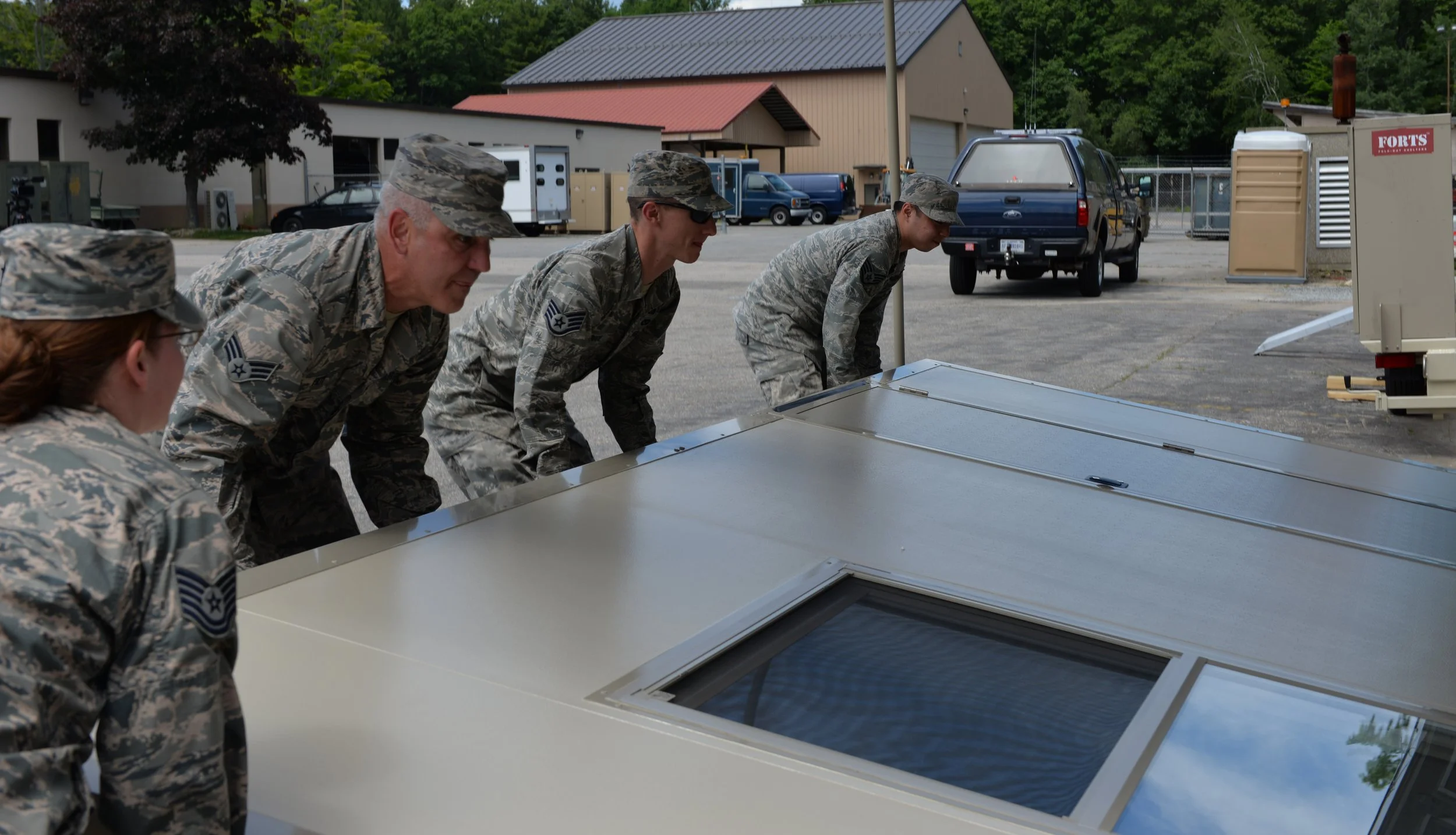 Military personnel in camouflage uniforms inspecting a solar panel on a shady outdoor setting with vehicles and buildings in the background.