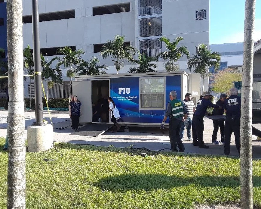 People unloading a mobile medical trailer labeled 'FIU Florida Advanced Surgical Transport Team' outside a building with palm trees.