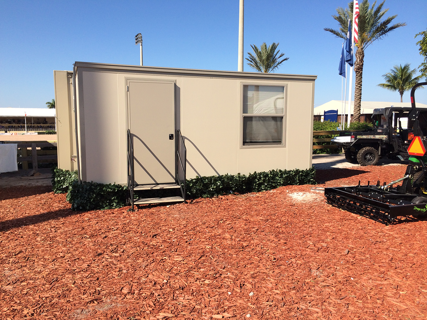 Small white prefabricated building with steps leading to a door and window, surrounded by red mulch and greenery, with palm trees, flags, and outdoor equipment in the background.