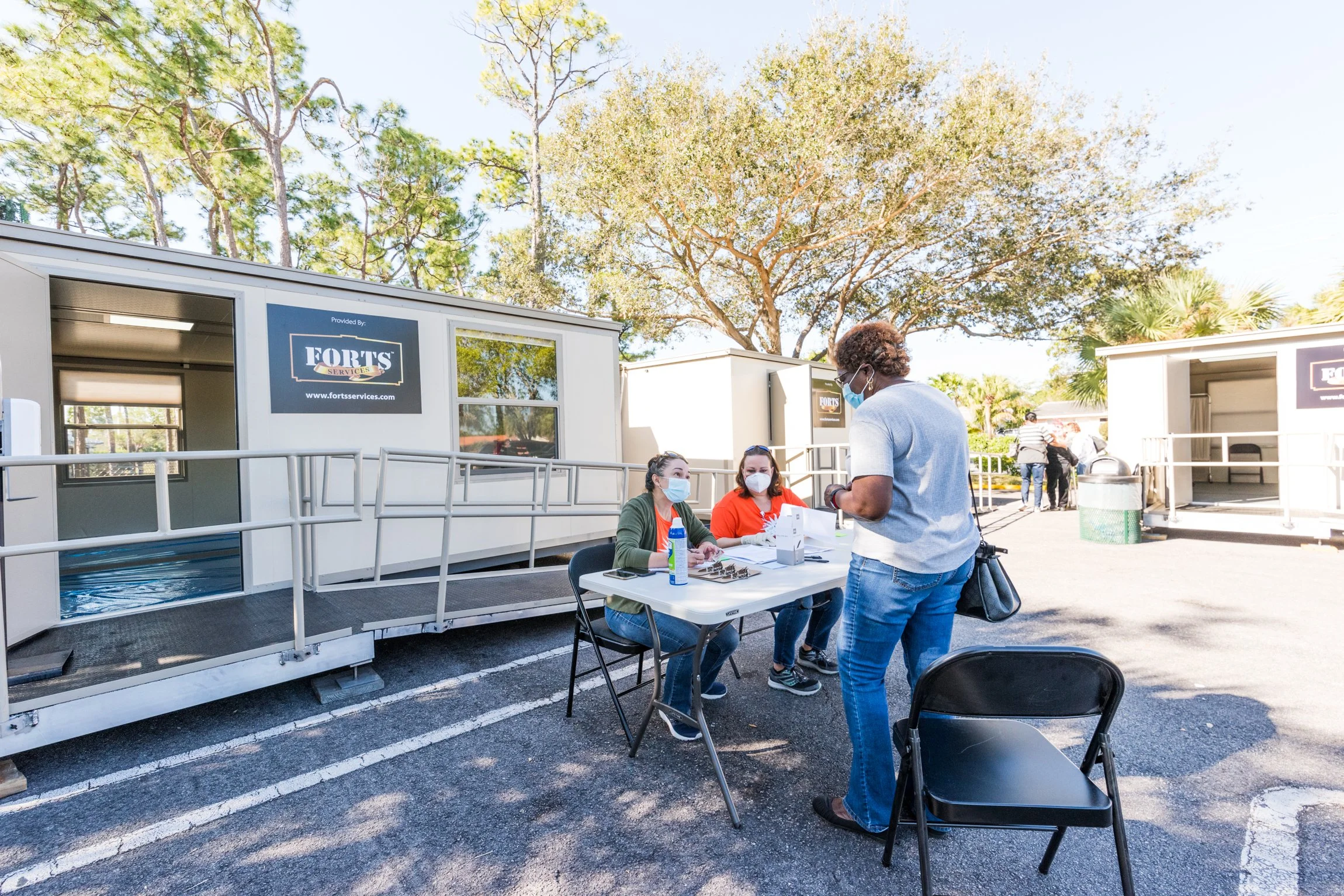 People wearing masks at a registration table outside next to mobile units with 'Fort's Services' logos, on a sunny day with trees in the background.