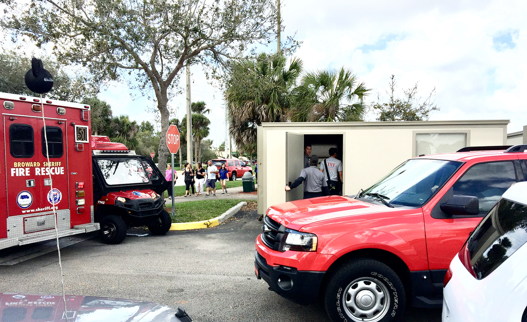 Emergency response vehicles parked near a small building with people walking nearby and some entering the building, including a fire rescue truck and a red utility vehicle.