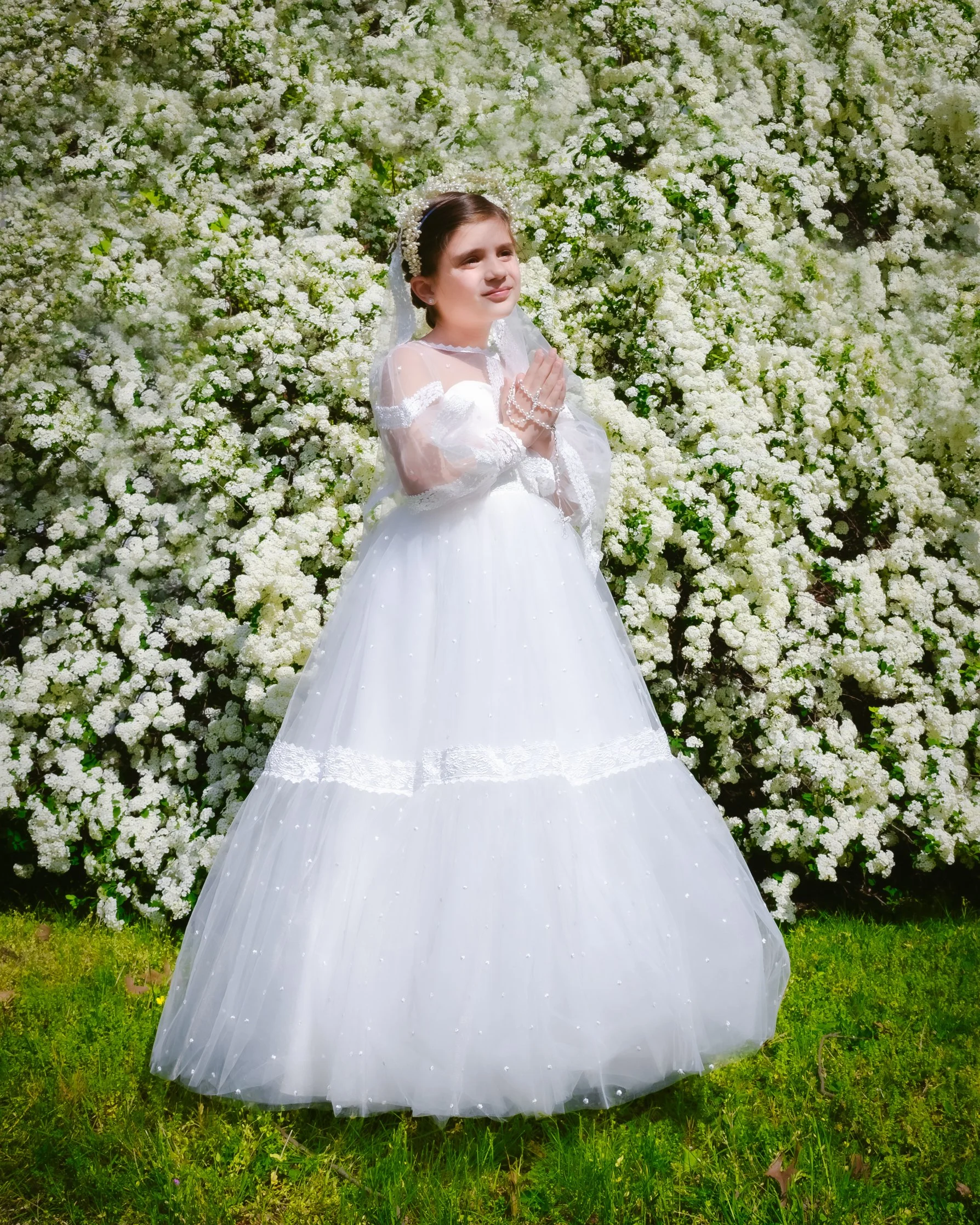 Young girl in a white communion dress with a veil and floral headband, standing in front of a flowering bush with white blossoms, holding her hands together.
