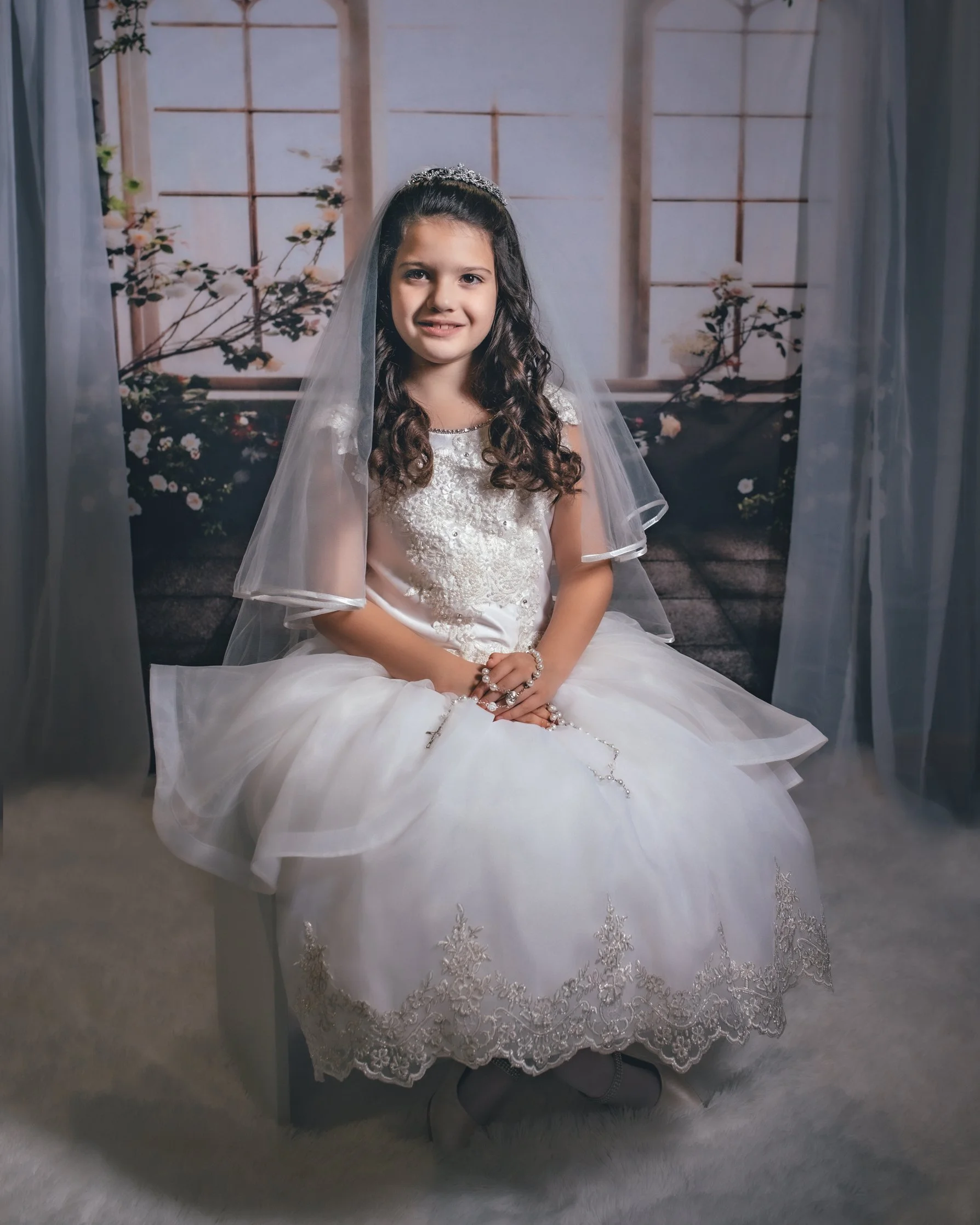 Young girl dressed in a white communion dress, wearing a tiara and veil, sitting on a chair with a prayer rosary in her hands, in front of a scenic backdrop of windows and flowers.