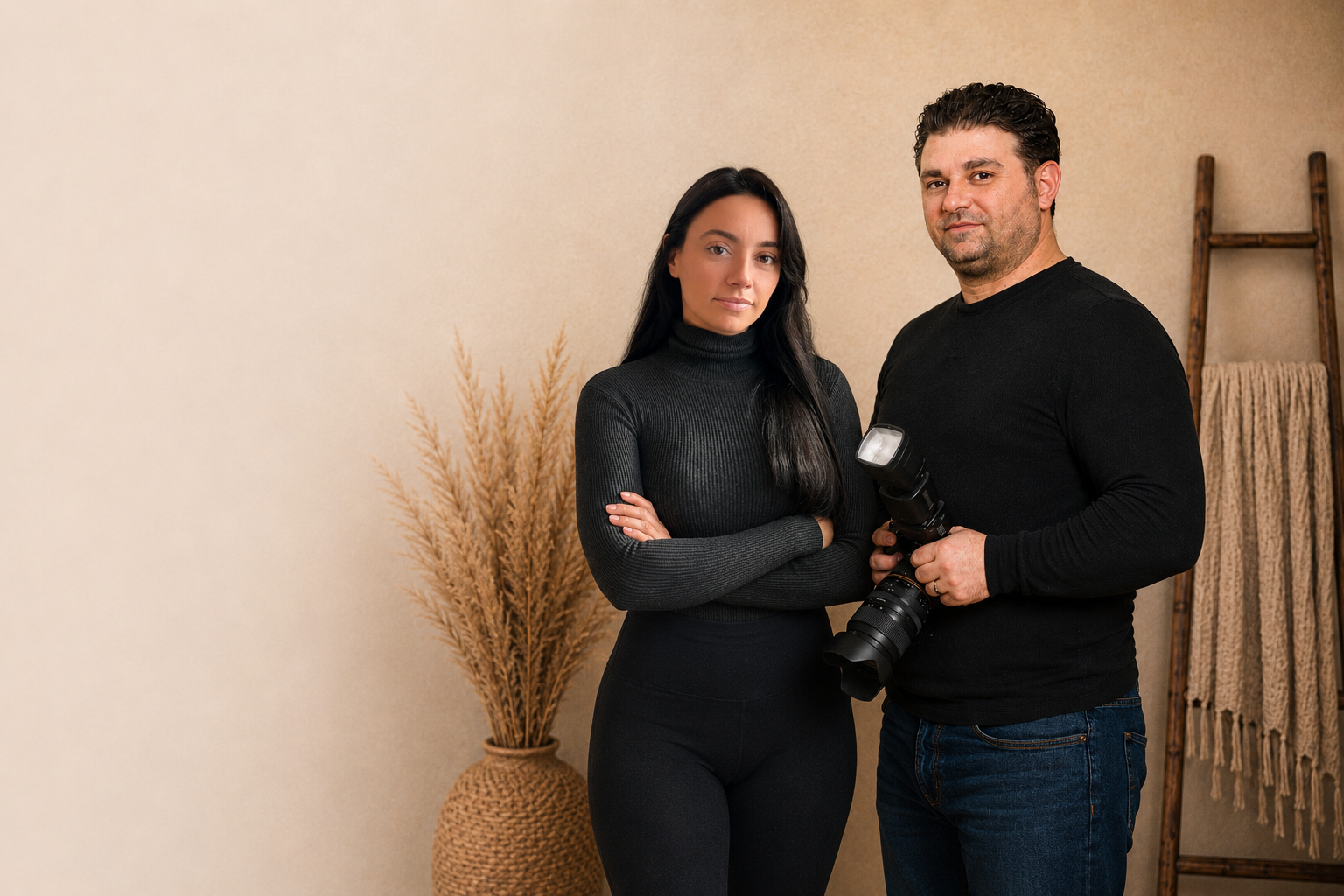 A woman and a man standing side by side in a beige room, with the woman crossing her arms and the man holding a professional camera with a flash attachment.