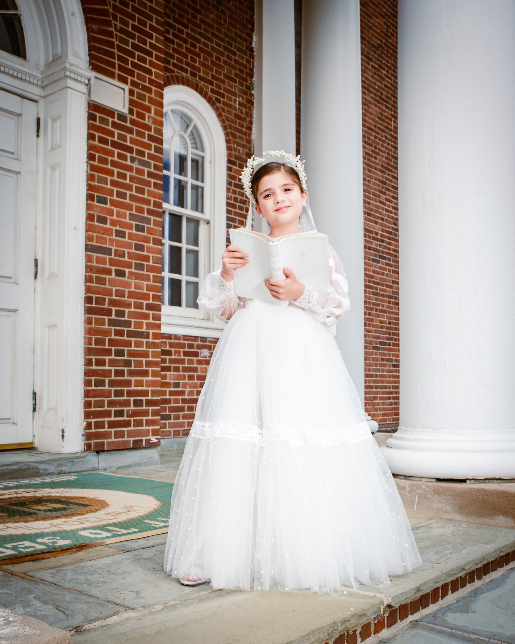 Young girl dressed in a white communion dress and veil, holding a book, standing outside in front of a brick building with white columns and windows.