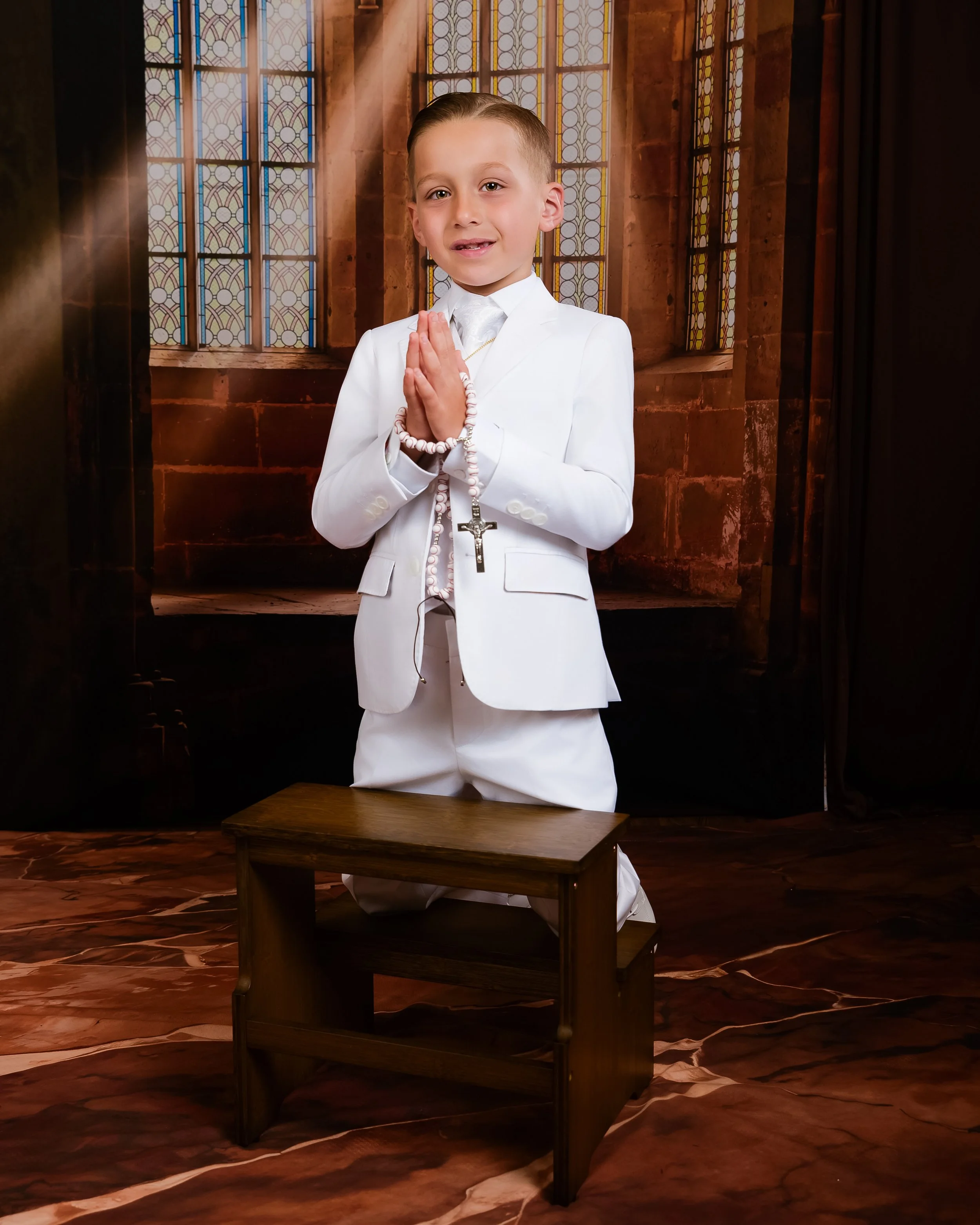A young boy dressed in a white communion suit kneeling on a wooden bench with a church background setting