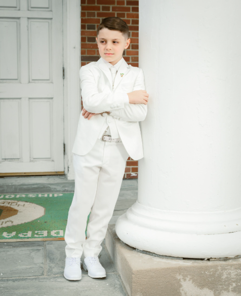 A young boy in a white suit standing outside next to a white pillar, with a brick wall and door in the background.