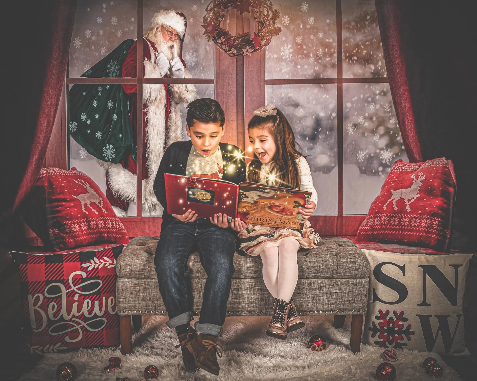 Two children, a boy and a girl, sitting on a couch in a cozy room decorated with Christmas décor, looking excitedly at a Christmas storybook with sparkles emitting from it. Santa Claus is seen outside the window, looking in, with a festive wreath han